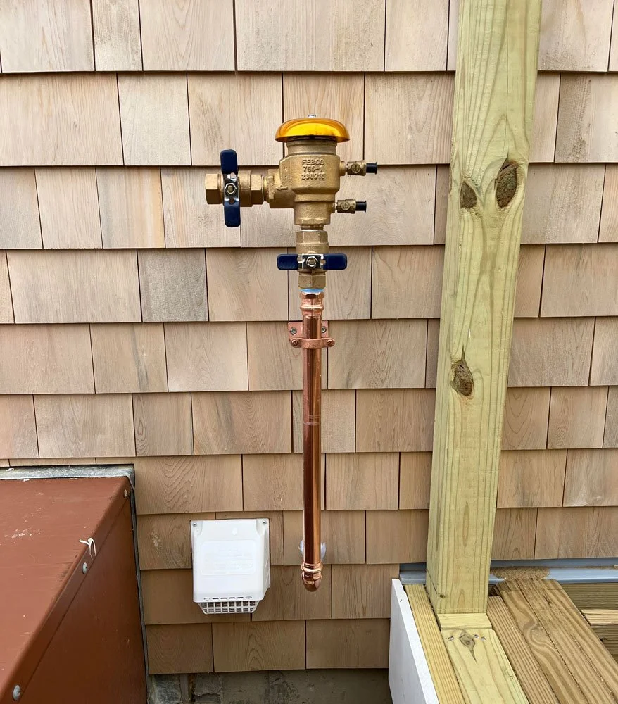 A plumbing fixture with a brass valve and a copper pipe attached to a wooden deck and shingled exterior wall.