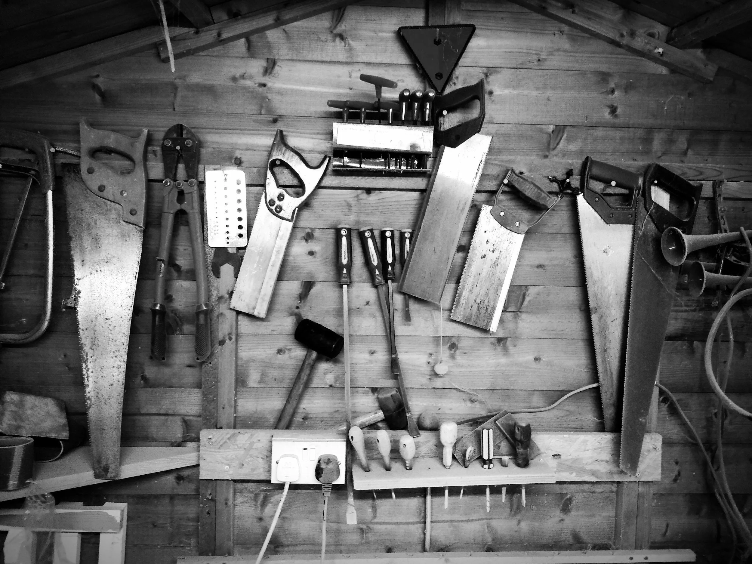 A black and white photo of various woodworking tools hanging on a wooden wall. Items include hand saws, clamps, screwdrivers, a triangular level, and an electrical outlet with plugs.