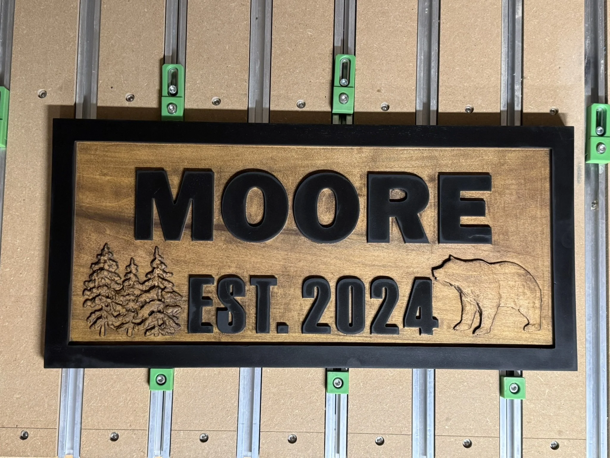 Close-up of a decorative wall hanging featuring the word 'MORE' in black letters on a wood background, surrounded by tree images and a mountain outline, mounted on a beige surface with green and silver mounting hardware.