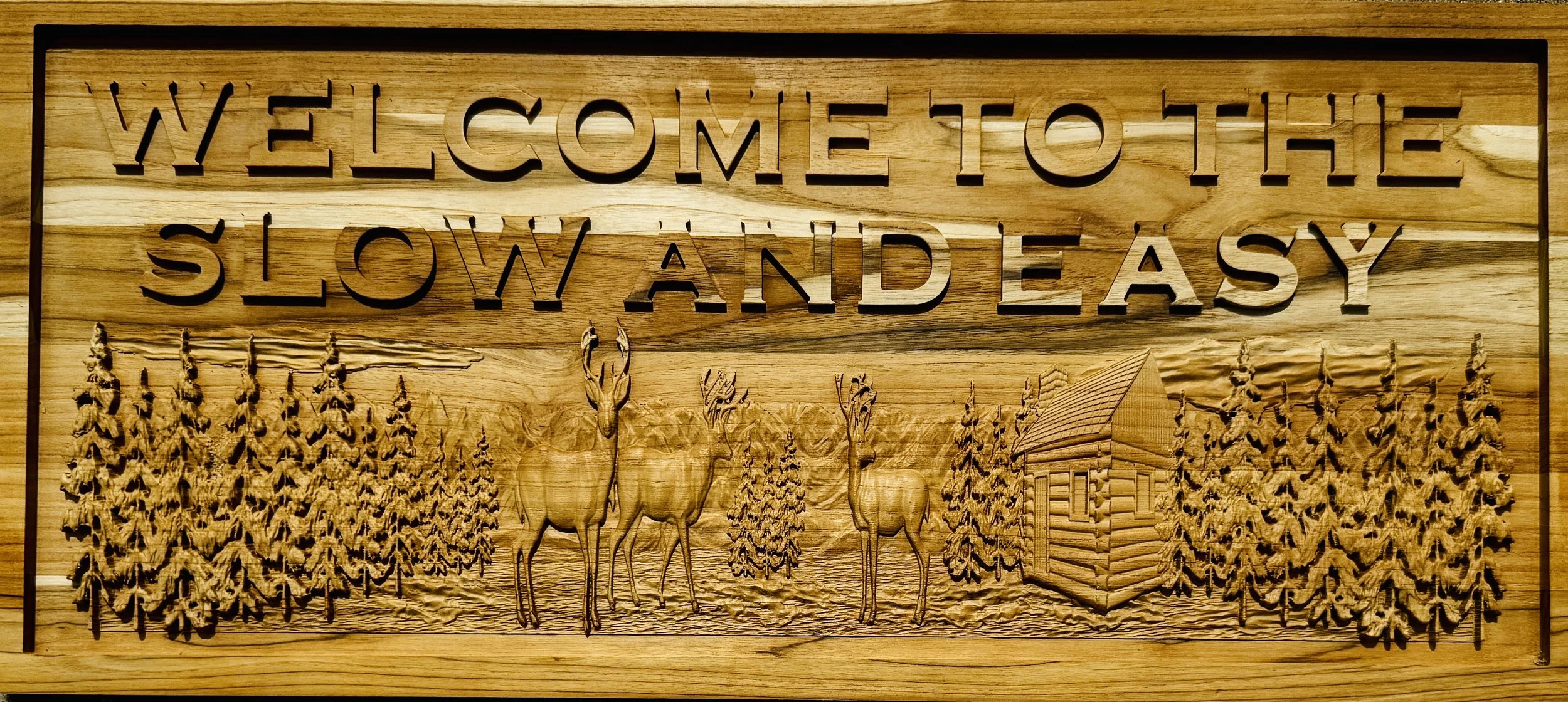 A wooden plaque with a carved scene of a forest, deer, and a small cabin, with the words "Welcome to the slow and easy" carved at the top.