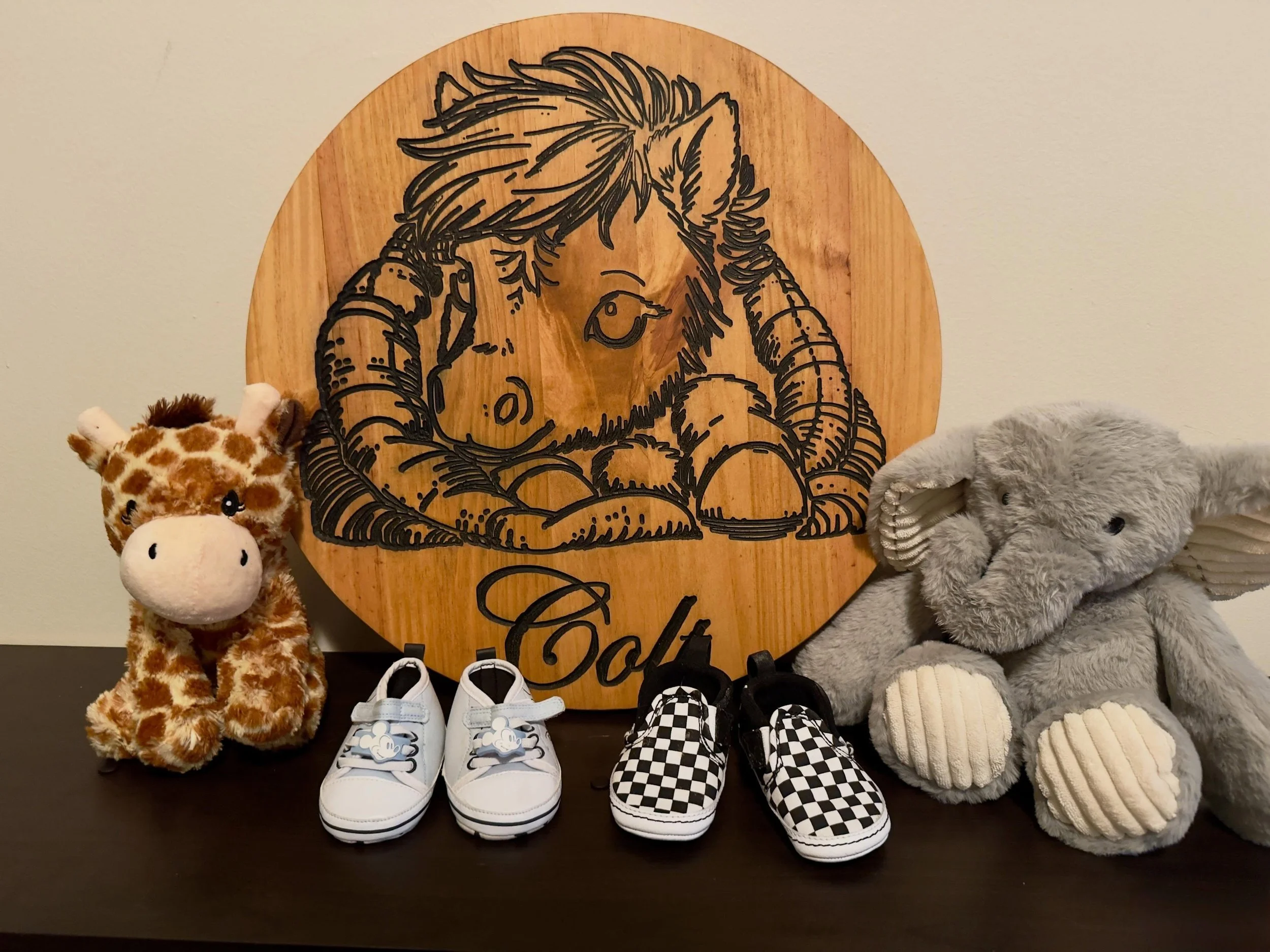 A display of children's shoes, plush toys (a giraffe and a gray bear), and a wooden art piece with a drawing of an astronaut and animal. The shoes include checkered slip-ons, sneakers, and knitted slippers.