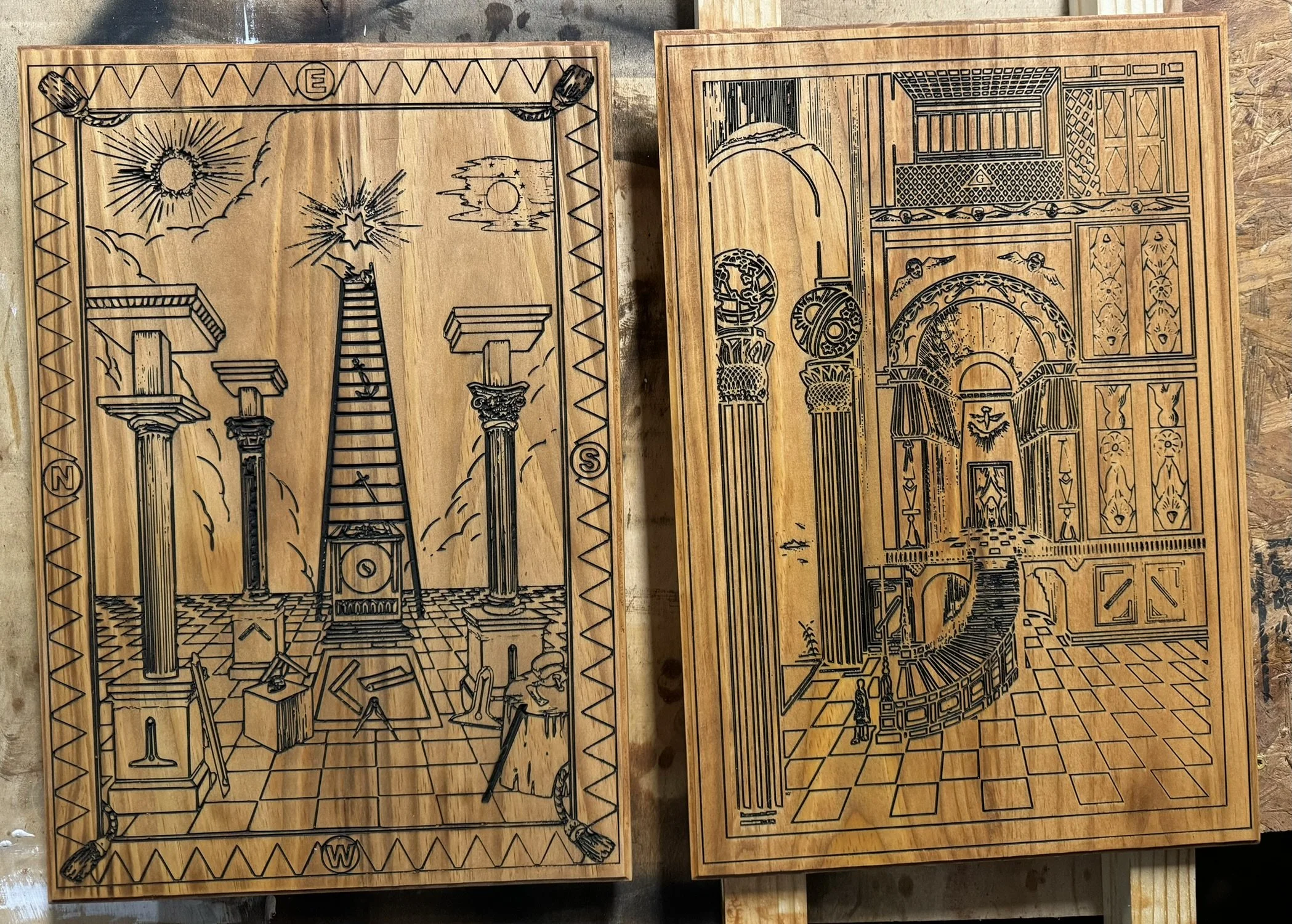 Two wooden boards with intricate black engraved designs. The left board features columns, a ladder, clouds, the sun, a star, and some symbols. The right board depicts architecture with doors, windows, arches, globes, and decorative patterns.
