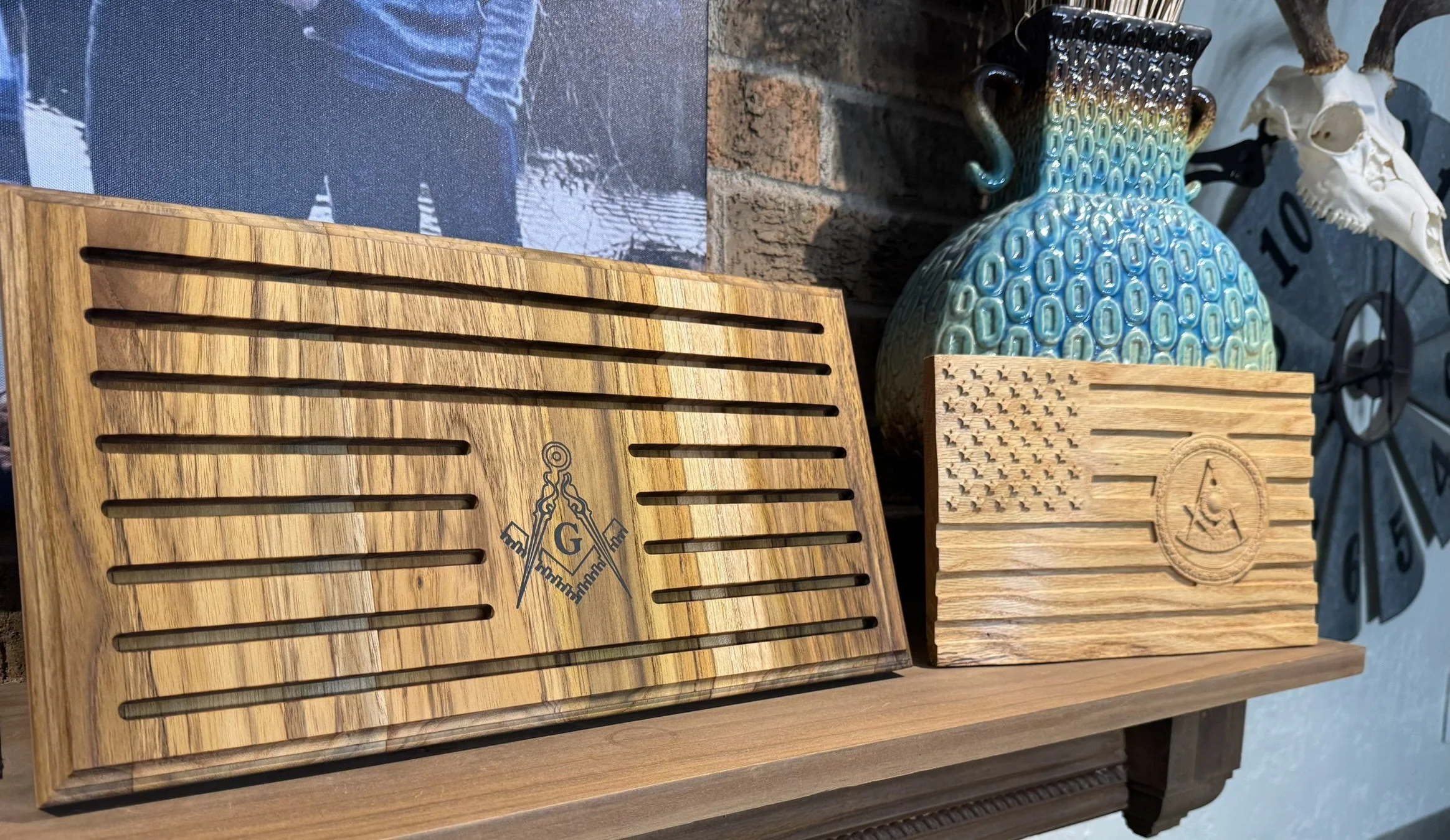 Two wooden boards with evenly spaced horizontal slots, one branded with a Freemason symbol, and the other with an encircled letter A. Behind them, decorative vases, one with a peacock feather design and the other a pattern of small stars, placed on a