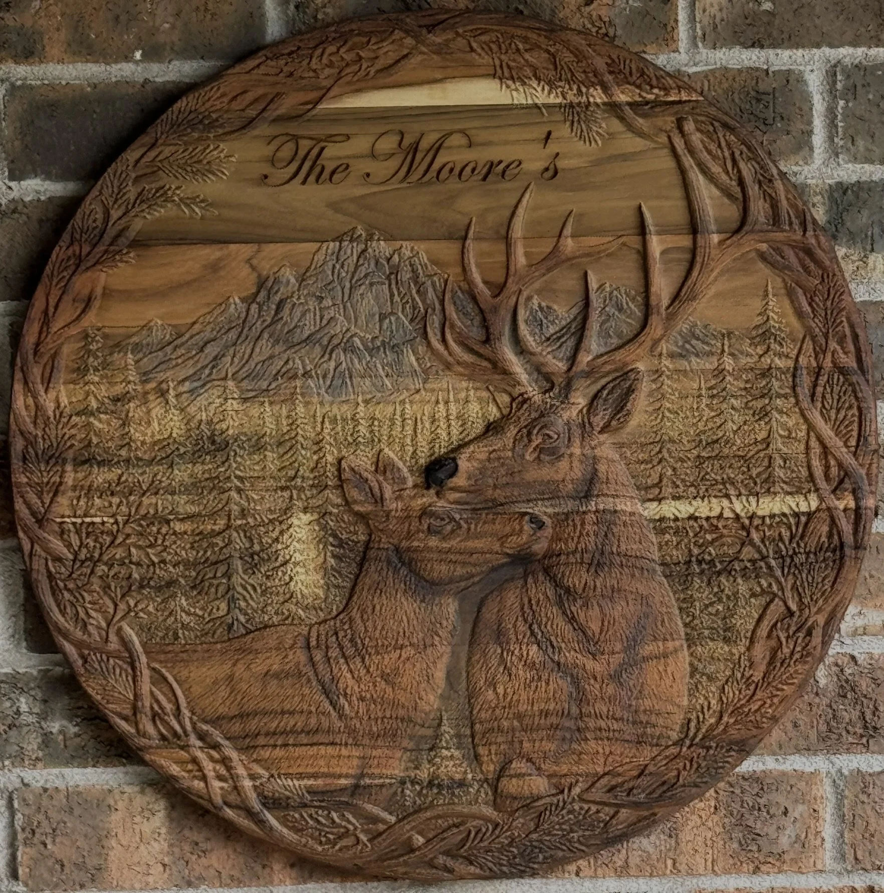 A round wooden plaque with carved mountain peaks, pine trees, and an elk with large antlers. The top of the plaque has the engraved text 'The Moore's' in cursive.