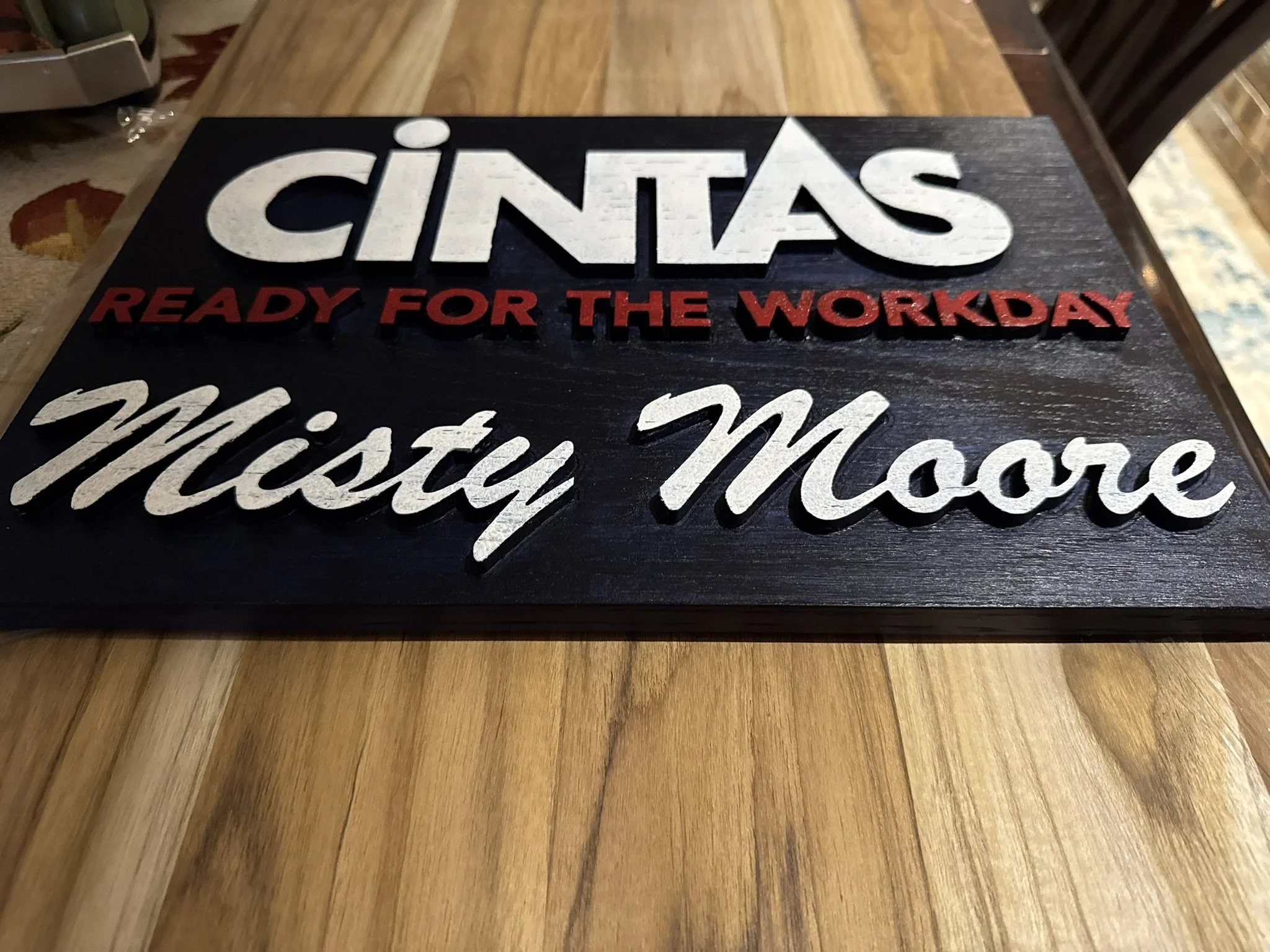 Close-up view of a decorative sign with the words "Cintas" in large white letters, "Ready for the Workday" in smaller red letters, and part of the phrase "Wichita, M..." in cursive. The sign appears to be made of wood with a dark background.