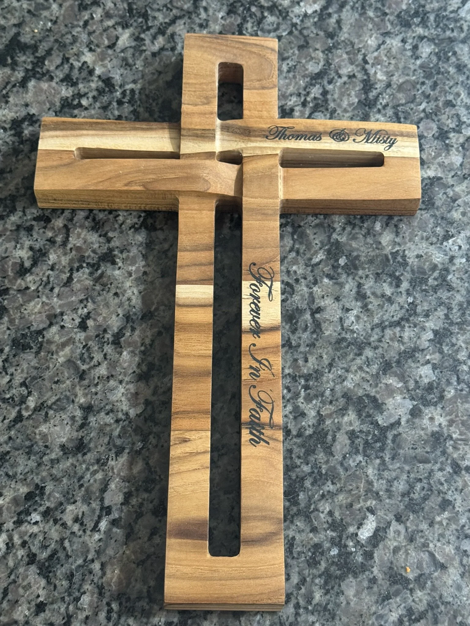 A wooden cross with engraved text 'Thomas & Misty' on the right side and partially visible text on the vertical part, placed on a dark granite countertop.