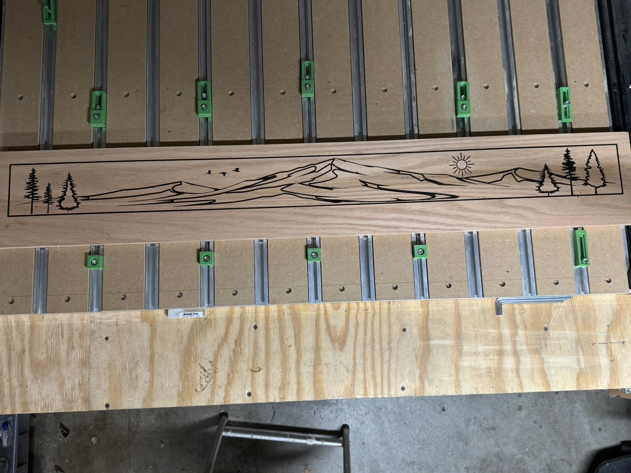 A wooden panel with a scenic mountain landscape engraved in black, showing mountains, trees, and birds, placed on a workbench.
