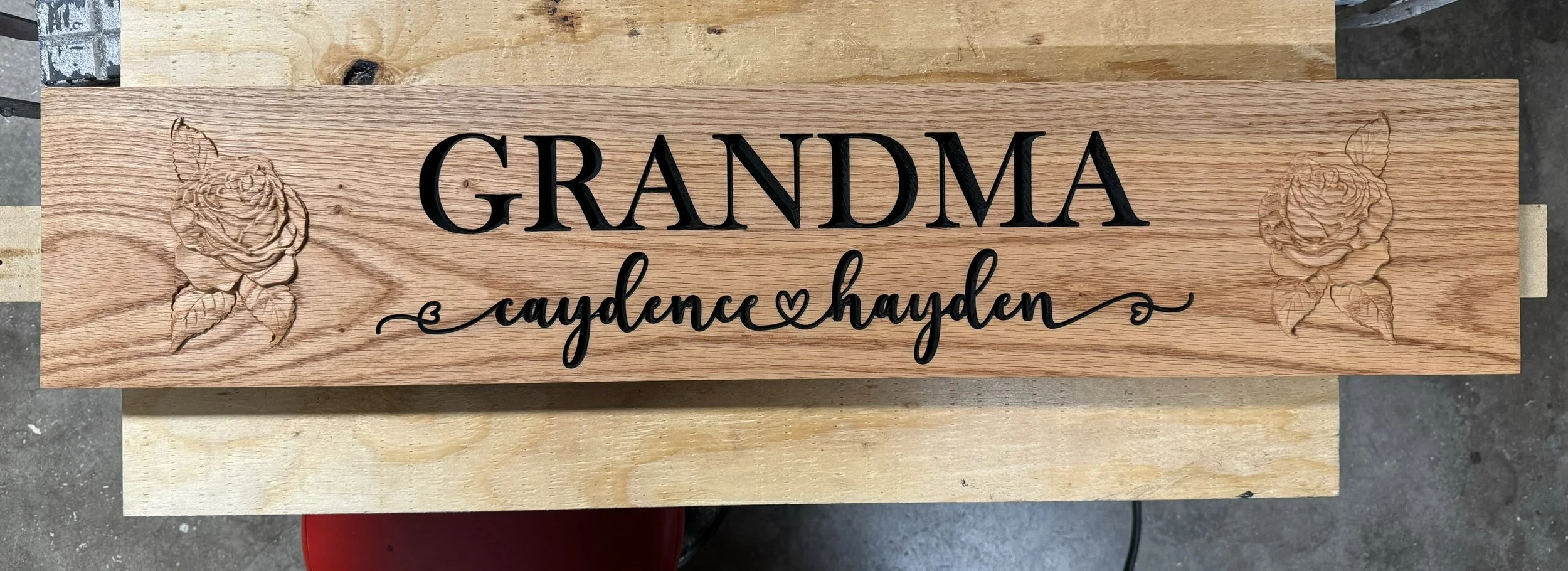 Wooden plaque with engraved roses on each end, black raised lettering that says 'Grandma', and cursive writing underneath that says 'Caydance & Hayden' with hearts on either side.