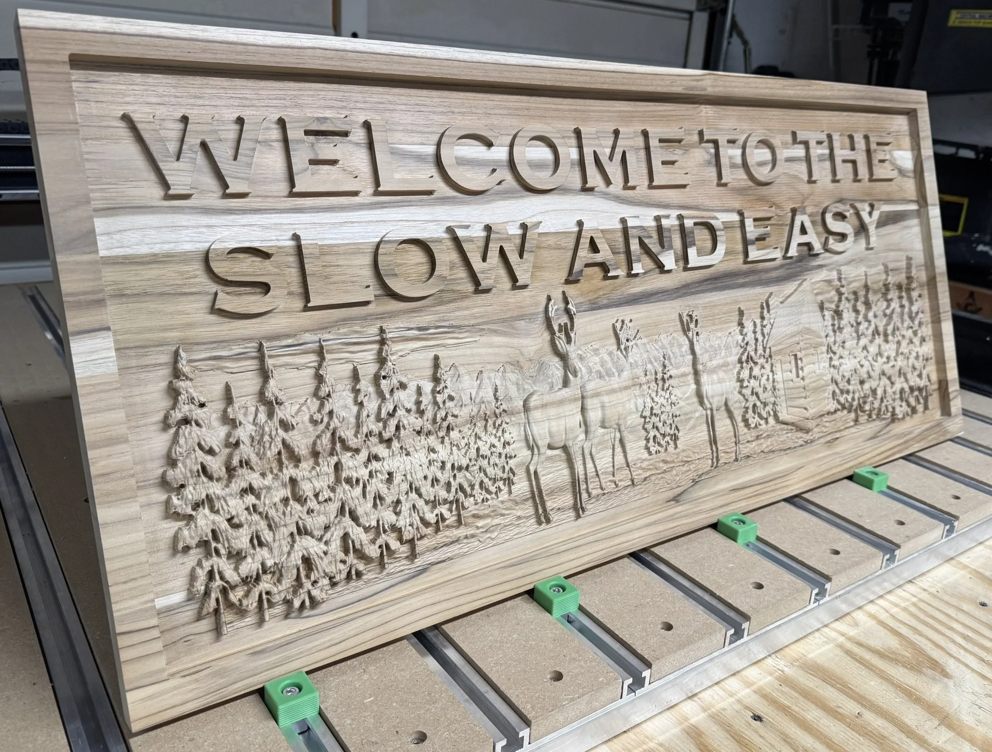 Wooden sign with engraved text and carved mountain trees, resting on a workbench in a workshop setting.