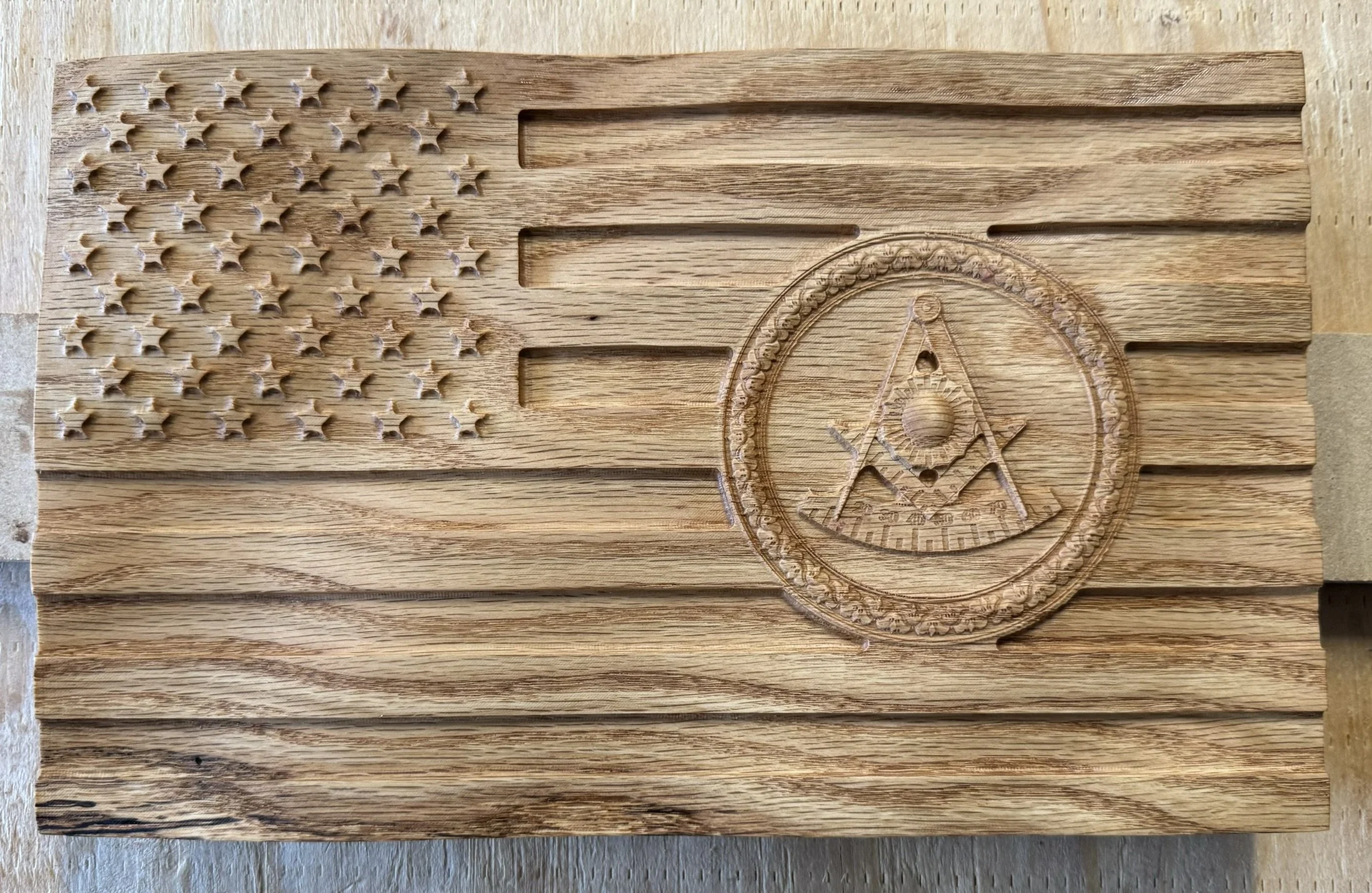 Wooden plaque designed in the style of the American flag with stars on the left and stripes on the right, featuring a carved emblem with an eye, a compass, and a graphic of layered mountain peaks.