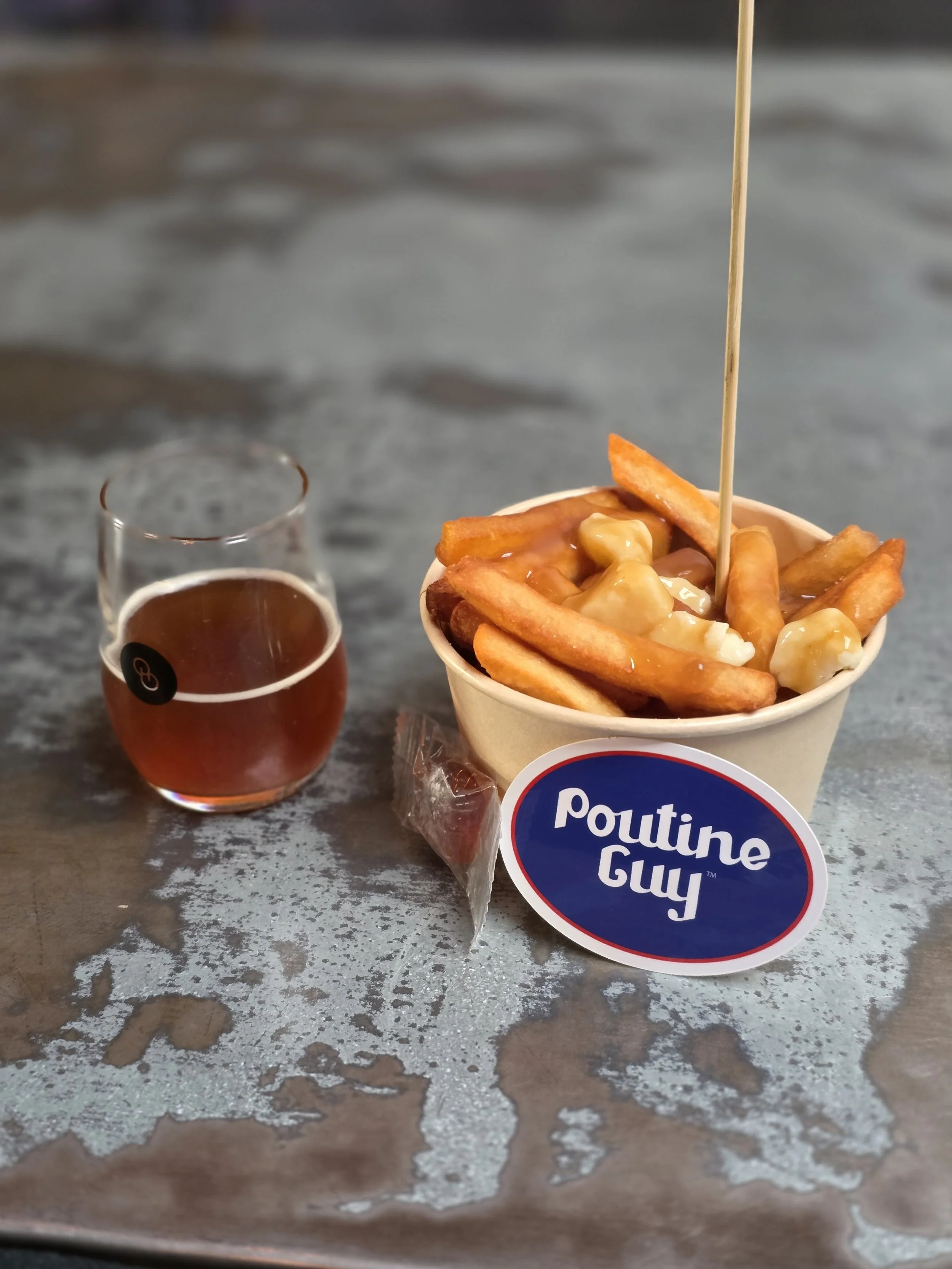 Cup of french fries topped with cheese and gravy, small glass of dark soda, on a weathered metal table, with a label reading "Poutine Guy."
