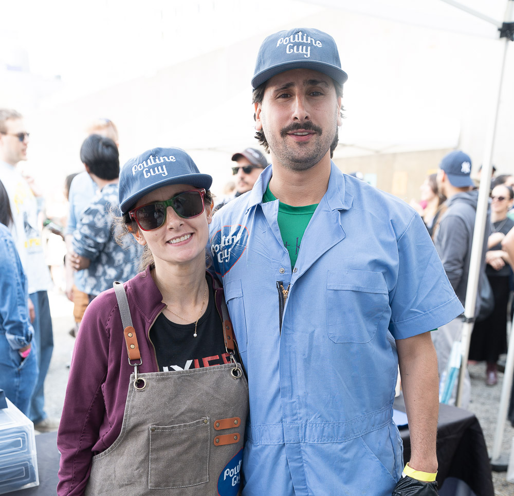 Two people wearing 'Poutine Guy' hats at an outdoor event, smiling for the camera. The woman is wearing sunglasses, a black shirt, and an apron. The man is dressed in a blue work shirt and cap, with many people in the background.