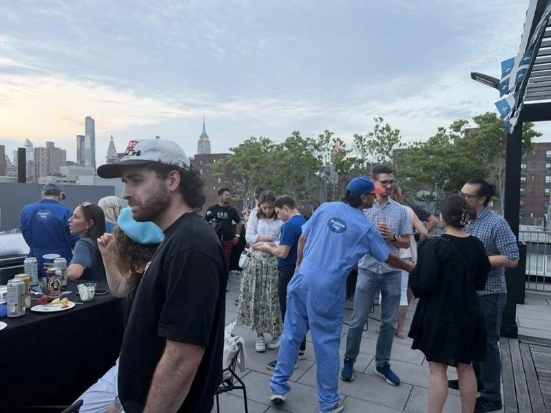 People socializing outdoors on a rooftop terrace with city skyline in the background, some holding drinks and others engaging in conversation.