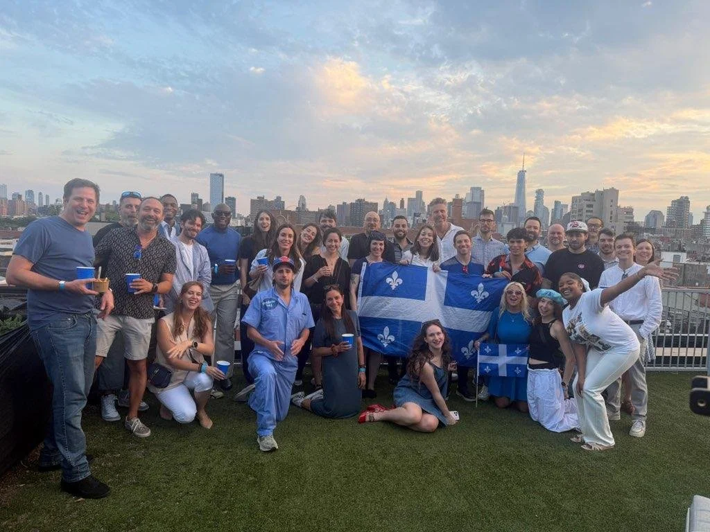 A group of people on a rooftop celebrating, holding two Quebec flags, with a city skyline during sunset in the background.