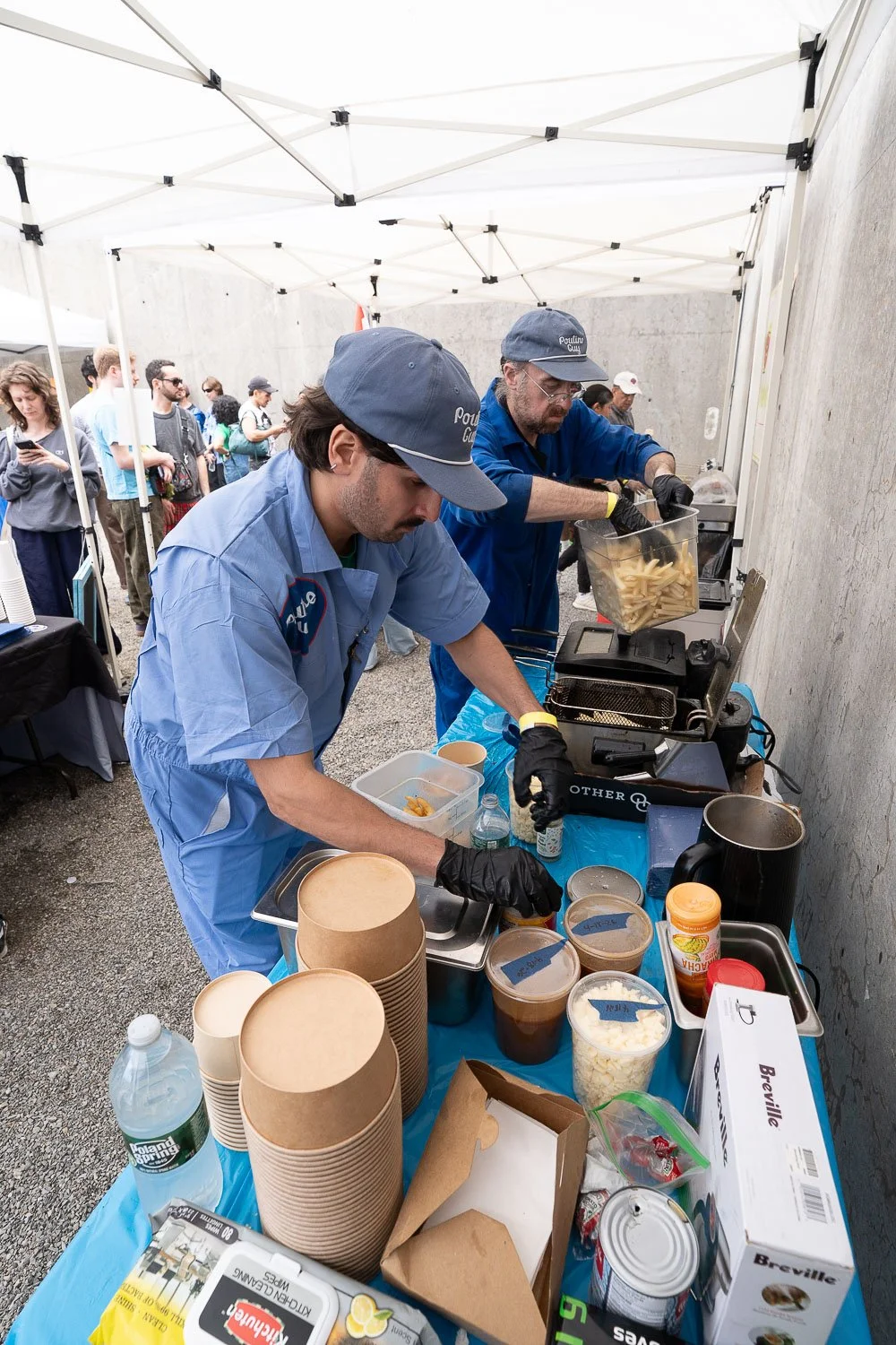 Two chefs in blue uniforms and black gloves preparing food at an outdoor event under a white canopy, with an audience waiting in line behind them.