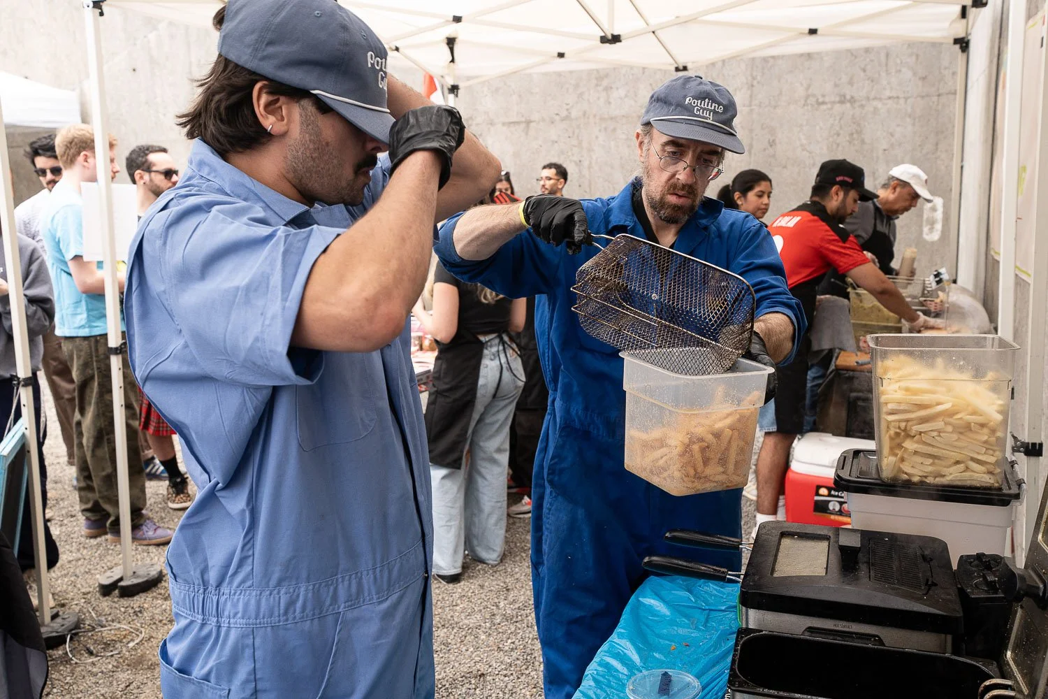 Two men in blue uniforms and hats labeled 'Poutine Guy' preparing poutine at an outdoor food stand, with a crowd of people in background.