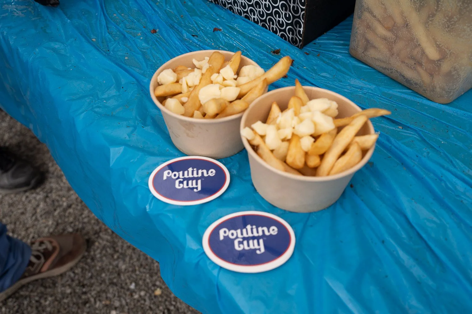 Two paper cups filled with French fries on a blue plastic tablecloth. There are two round stickers with white and red borders that say 'Poutine Guy' near the cups. In the background, part of a black box with white patterns and a large container is visible.
