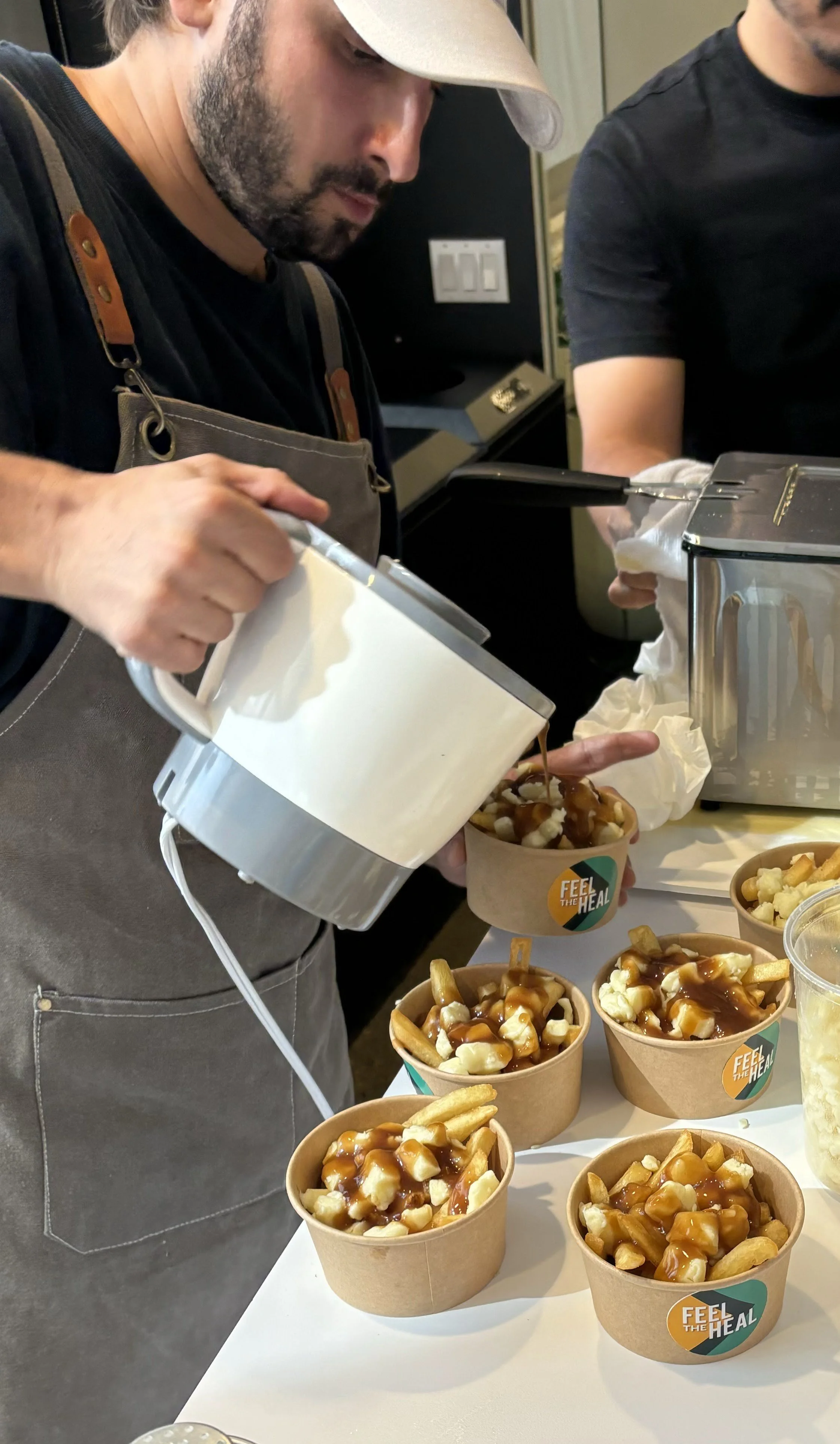 A person wearing a gray apron and beige cap is pouring caramel sauce from a metal pitcher onto cups of loaded poutine, which contains French fries, cheese curds, and gravy, on a white table. Several other cups of poutine are visible on the table.