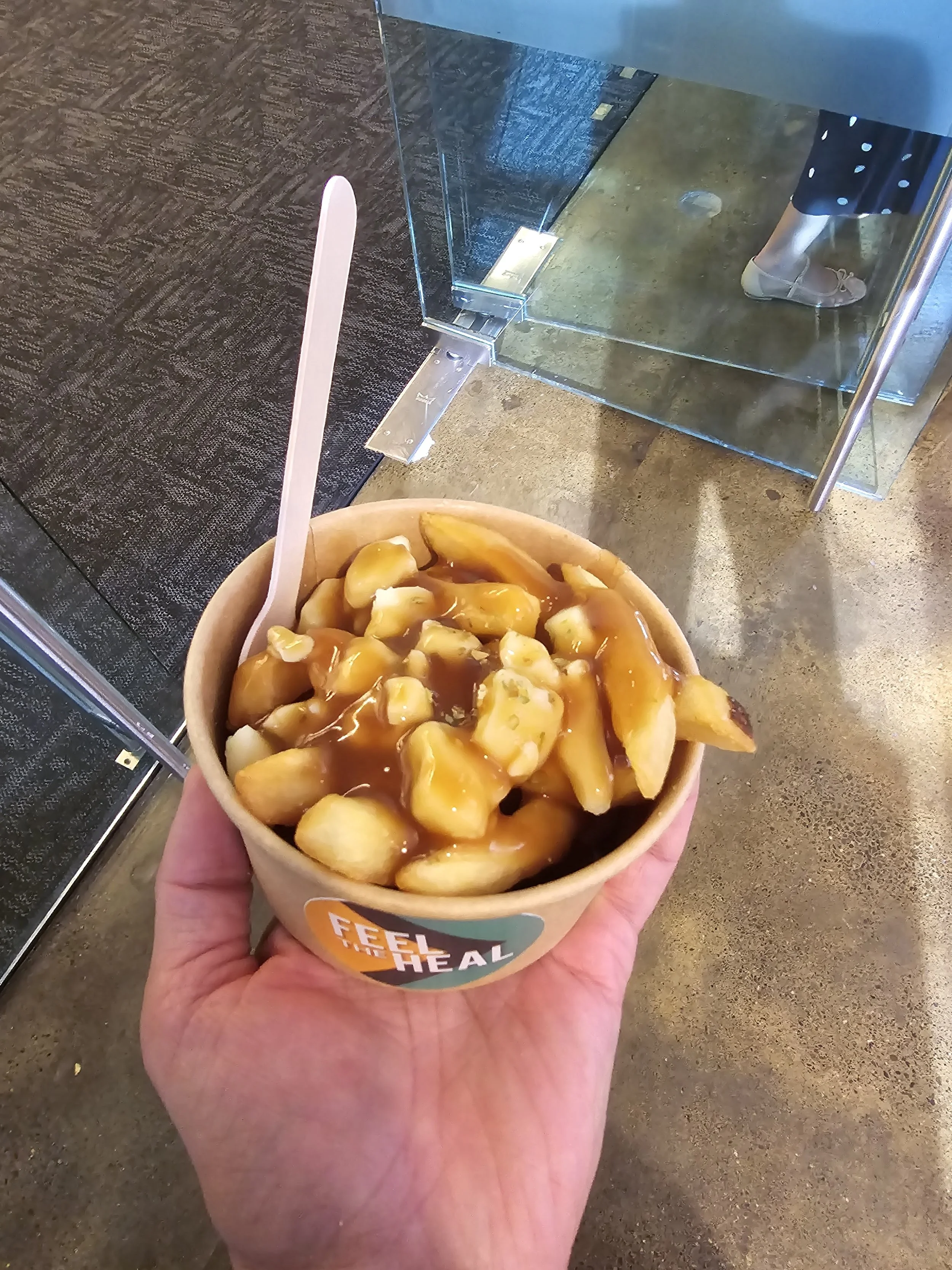 A person holding a bowl of peaches topped with caramel and other toppings in front of a reflective glass display case, with part of a person's foot and shoe visible in the background.