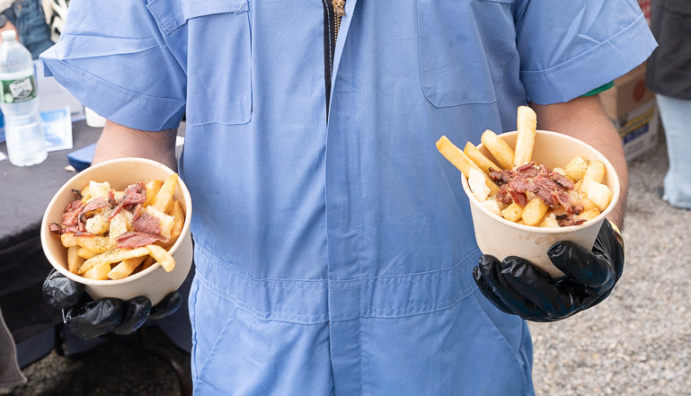 Person in blue uniform and black gloves holding two bowls of poutine with French fries, cheese curds, and gravy.