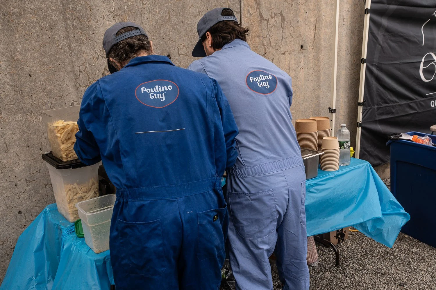 Two workers in blue uniforms with "poutine Guy" logos preparing food at a stand, with stacked paper cups, a water bottle, and food containers on the table.