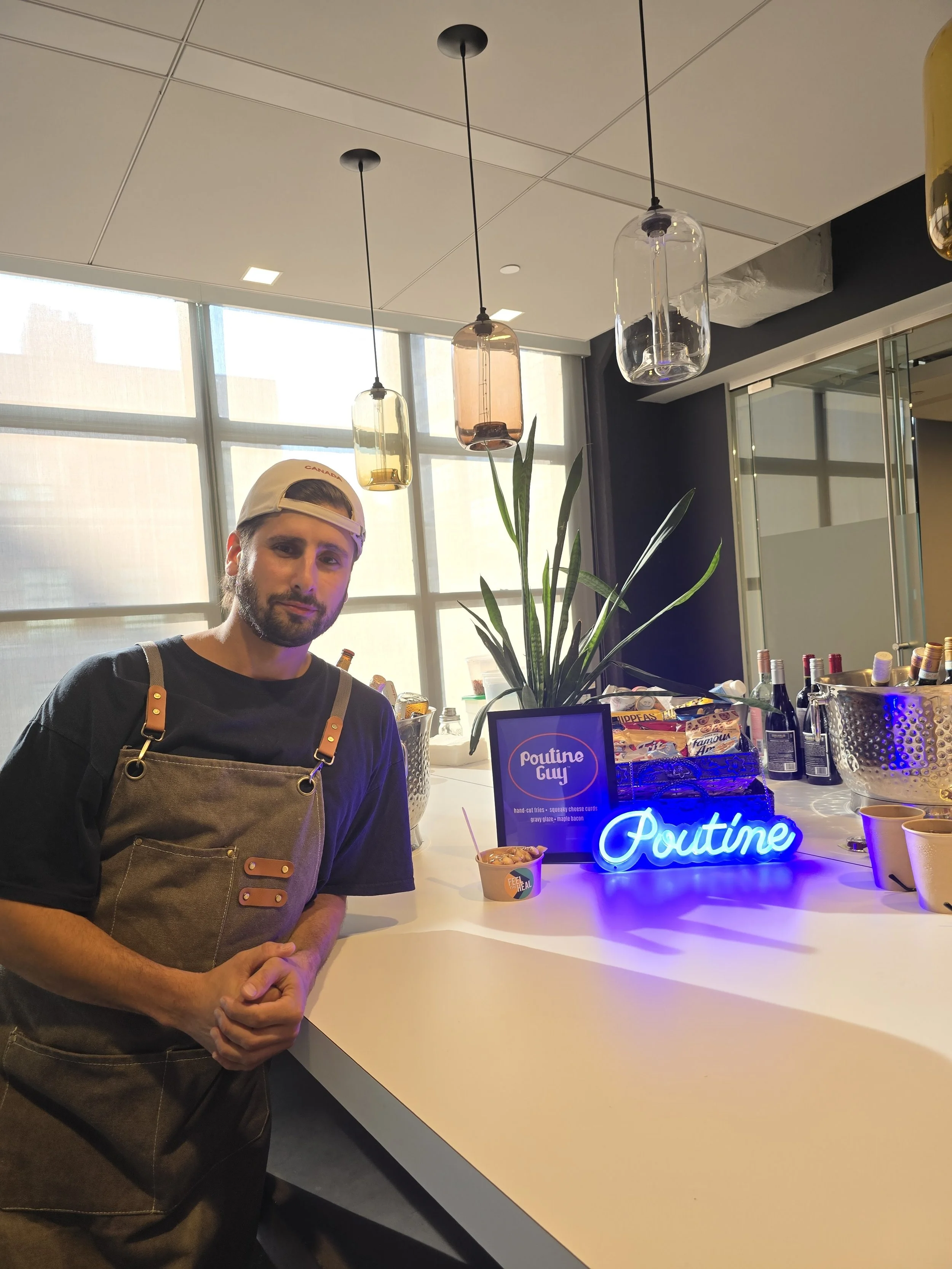 A young man with a beard and wearing a cream baseball cap and an apron stands at a white counter in a bright modern space with large windows. There are pendant lights hanging from the ceiling and a large plant behind him. On the counter are a sign reading 'Poutine Guy', a small container of poutine, various bottles of wine, and a neon sign that says 'Poutine' in bright blue.