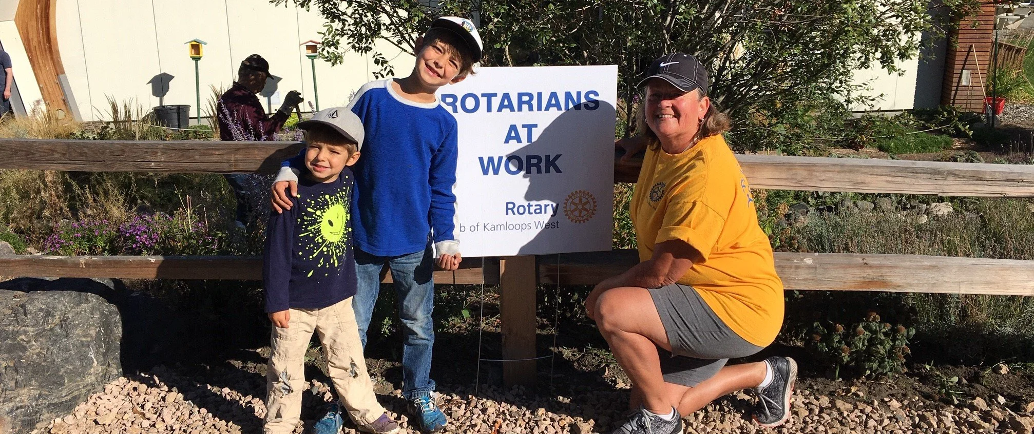 Three people posing outdoors in front of a sign that says "Rotarians at Work" with a Rotary International logo. There are two children, one adult woman, and a background with plants, trees, and a building.
