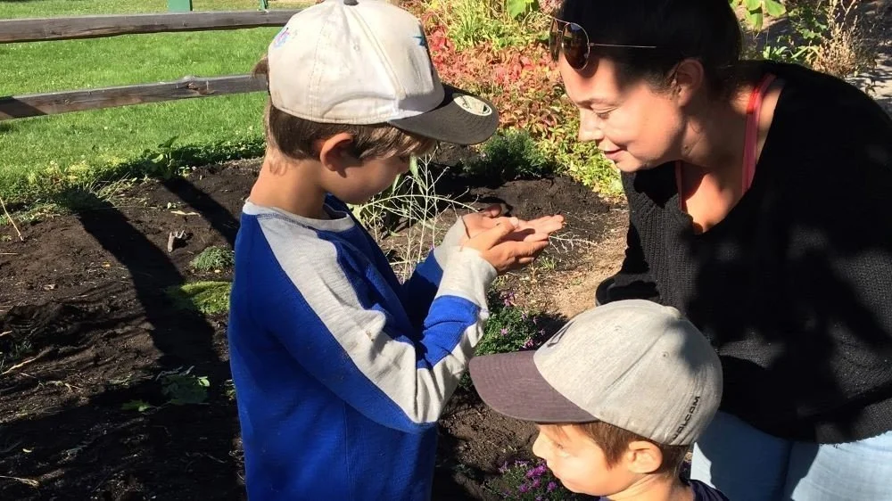 A woman and two children in a garden, with the woman assisting one of the children as he holds something in his hand and looks at it.