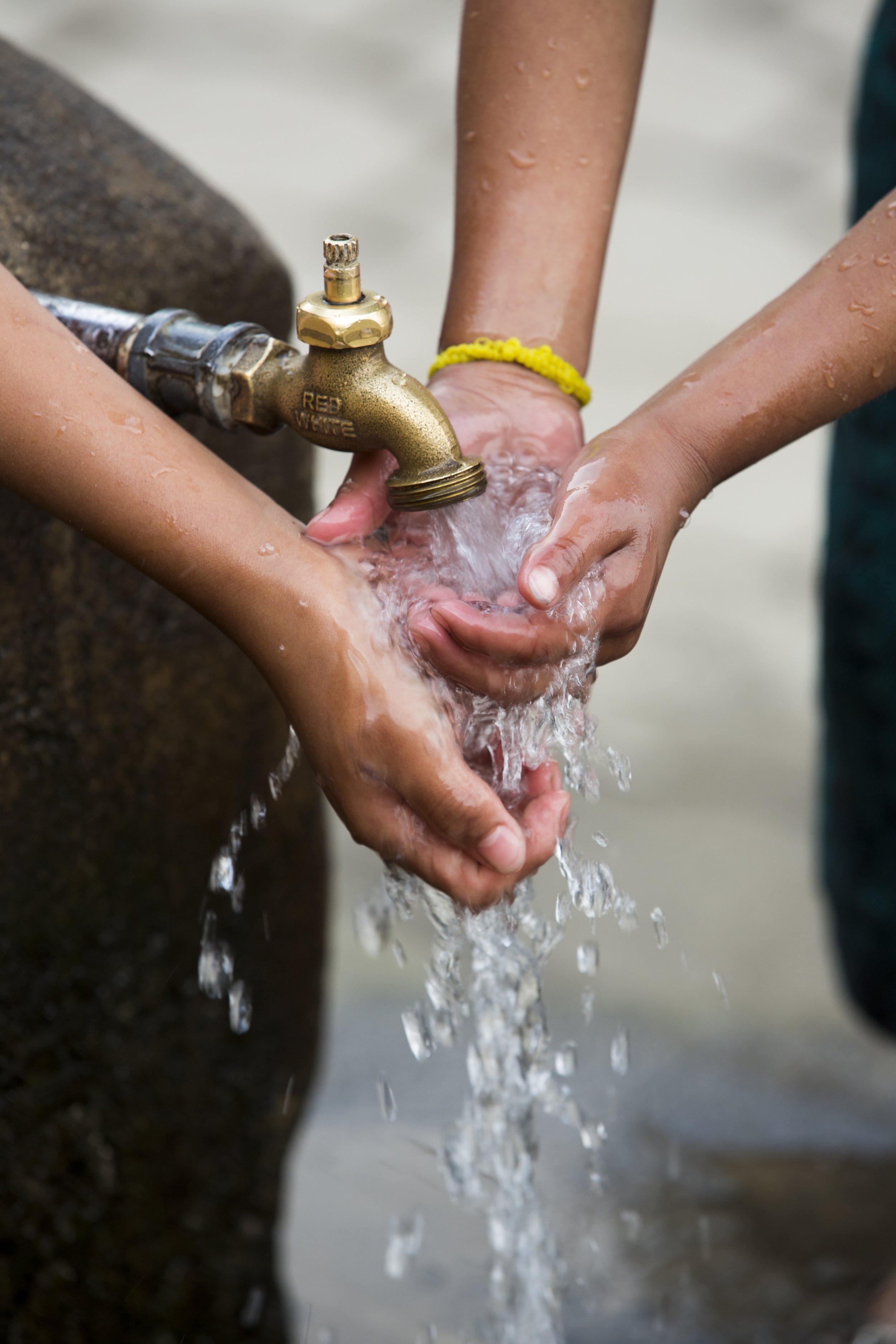 Two people are washing their hands together under a stream of water from an outdoor faucet. One person's hand is under water while the other's hand supports it. Droplets of water and wet skin are visible.