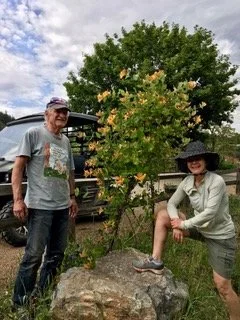 Two people, a man and a woman, are outdoors near a large tree and a rock. The man is standing on the left, wearing a cap, t-shirt, and jeans, while the woman is sitting on the rock, wearing a hat, long-sleeve shirt, and shorts. There is a vehicle and greenery in the background.