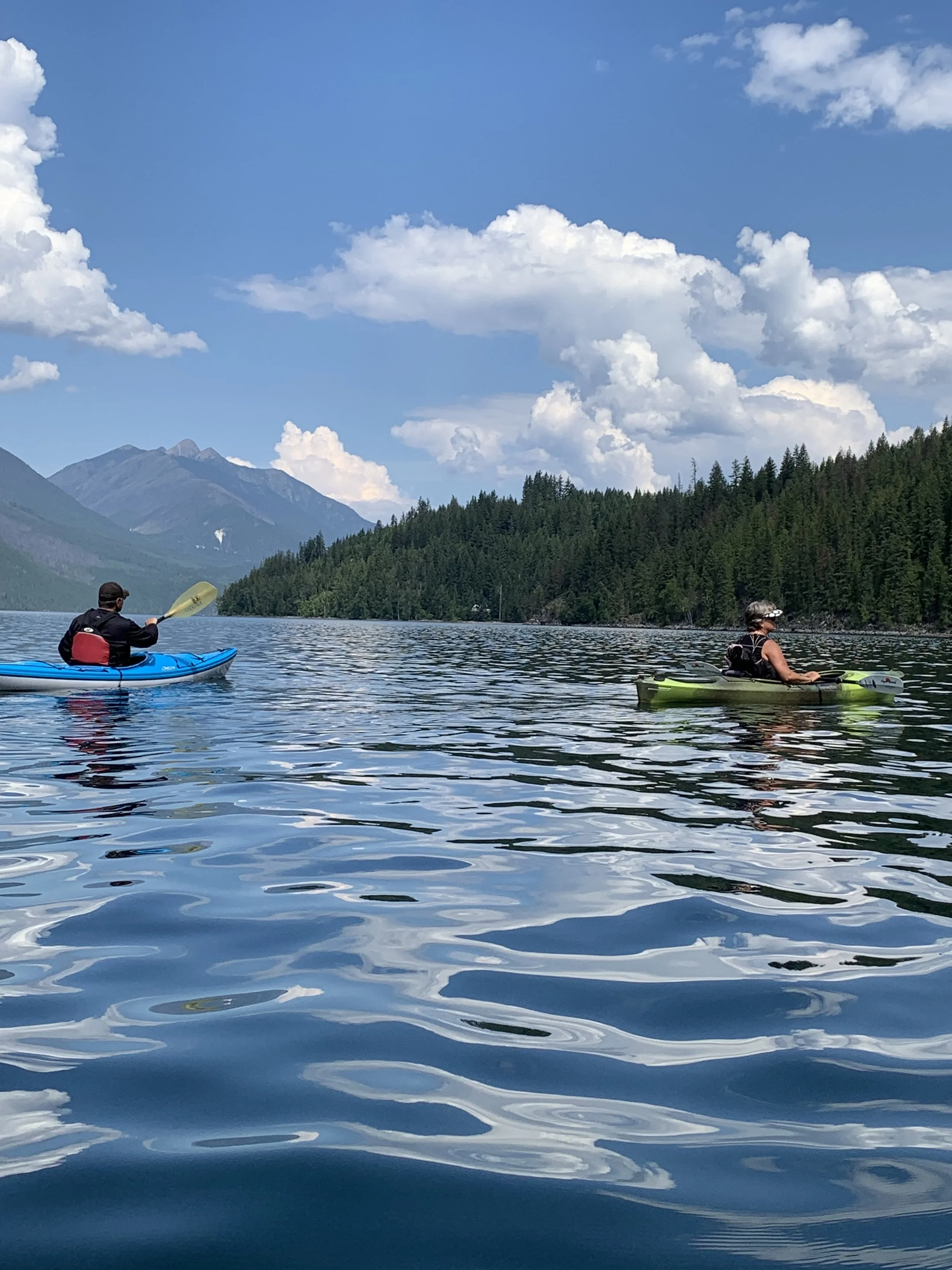 Two people kayaking on a calm lake surrounded by forested hills and mountains under a partly cloudy blue sky.
