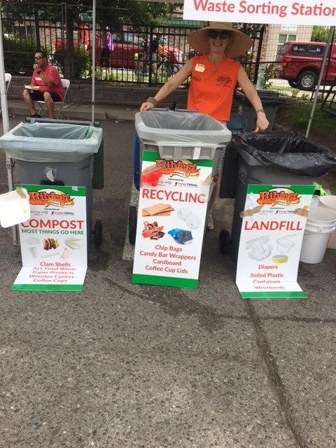 A woman standing behind three recycling bins labeled for compost, recycling, and landfill, at a waste sorting station. She is wearing a wide-brimmed hat and orange shirt, with others sitting and walking in the background.