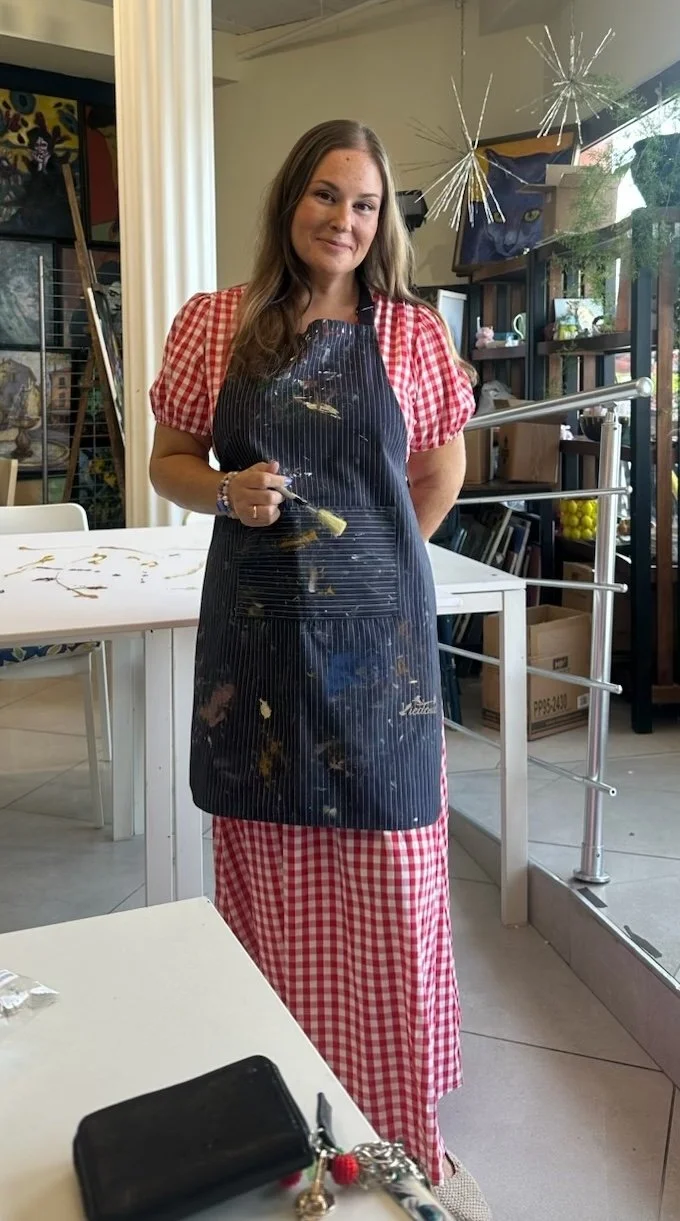 A woman wearing a black apron covered with paint stands in an art studio, holding a paintbrush. She is smiling and has long, wavy hair. The studio has shelves with art supplies, artwork, and a window with natural light.