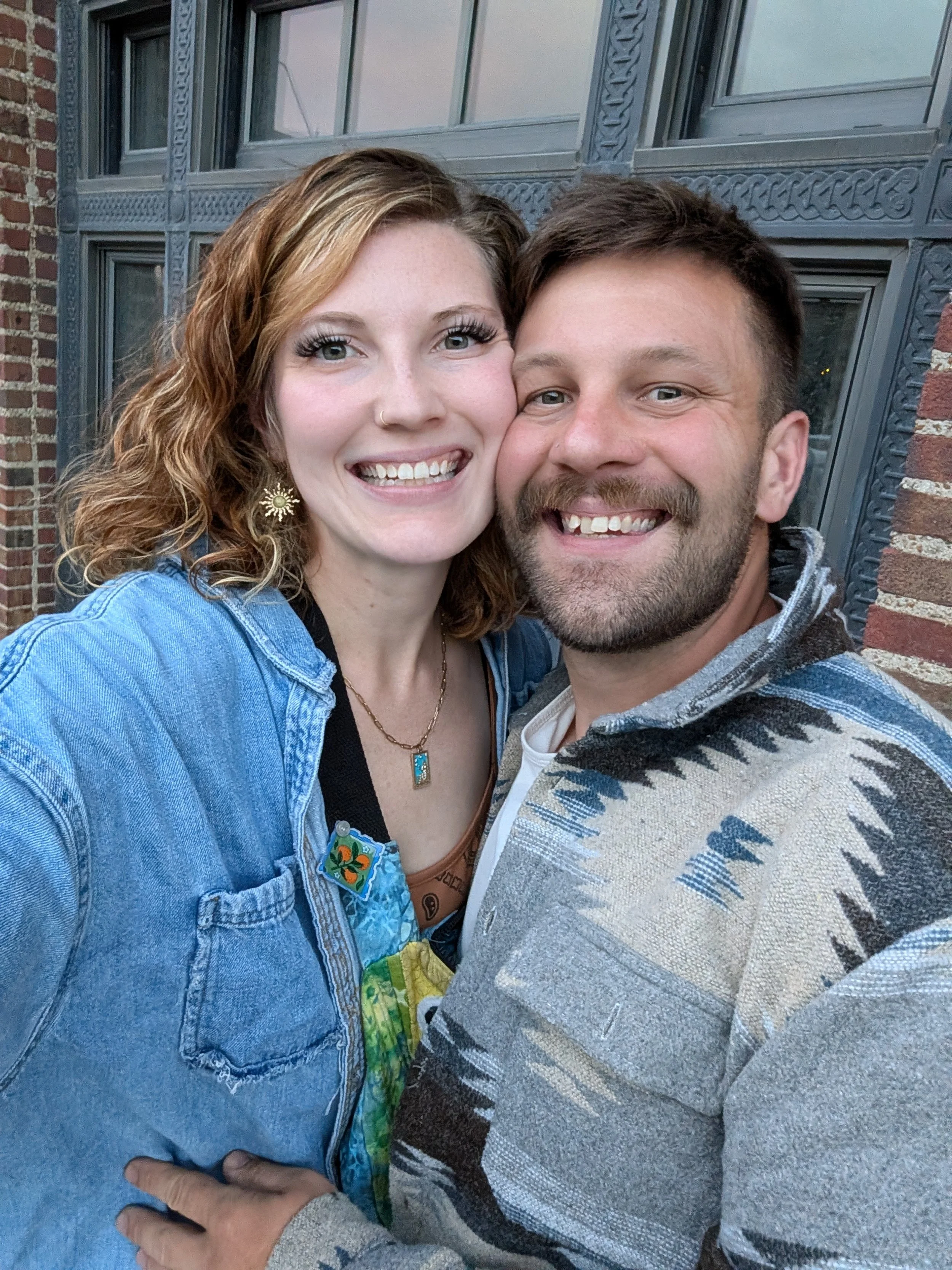 A smiling couple takes a selfie outside in front of a brick building with decorative blue-gray window frames. The woman has curly light brown hair, wears a denim jacket and gold earrings, and is dressed in colorful clothing. The man has short dark hair, a beard, and wears a striped sweater with a light shirt underneath, with his arm around the woman.