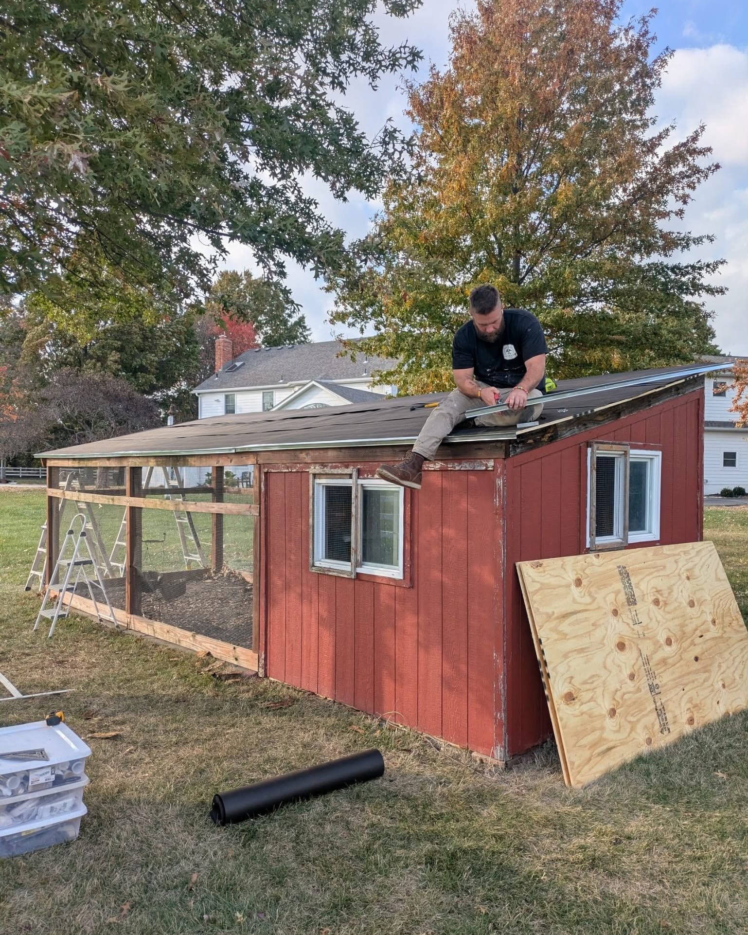 A man working on the roof of a small red wooden shed using a tool. The shed has windows and a porch, and trees with autumn foliage are in the background.
