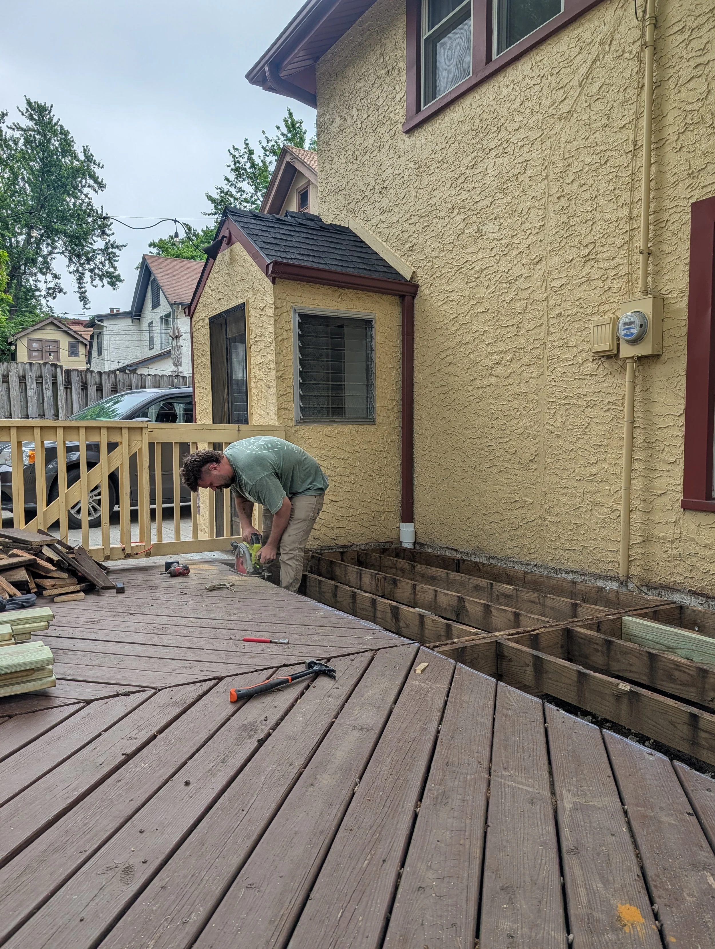 A man working on a wooden deck in a backyard, with part of the deck removed, and tools scattered around.