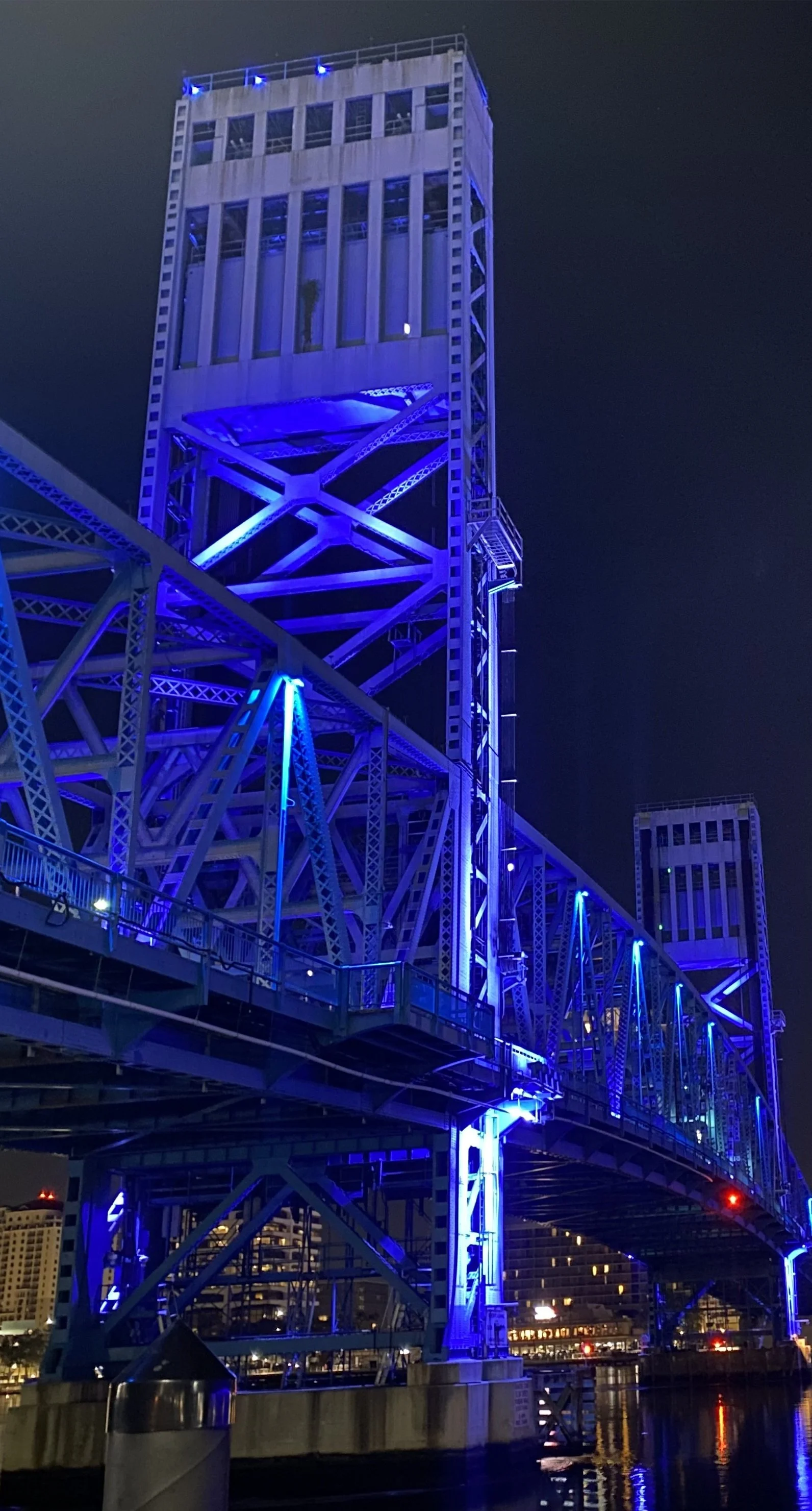Night view of a bridge illuminated with blue lights, with a tall building in the background and water reflecting the lights.