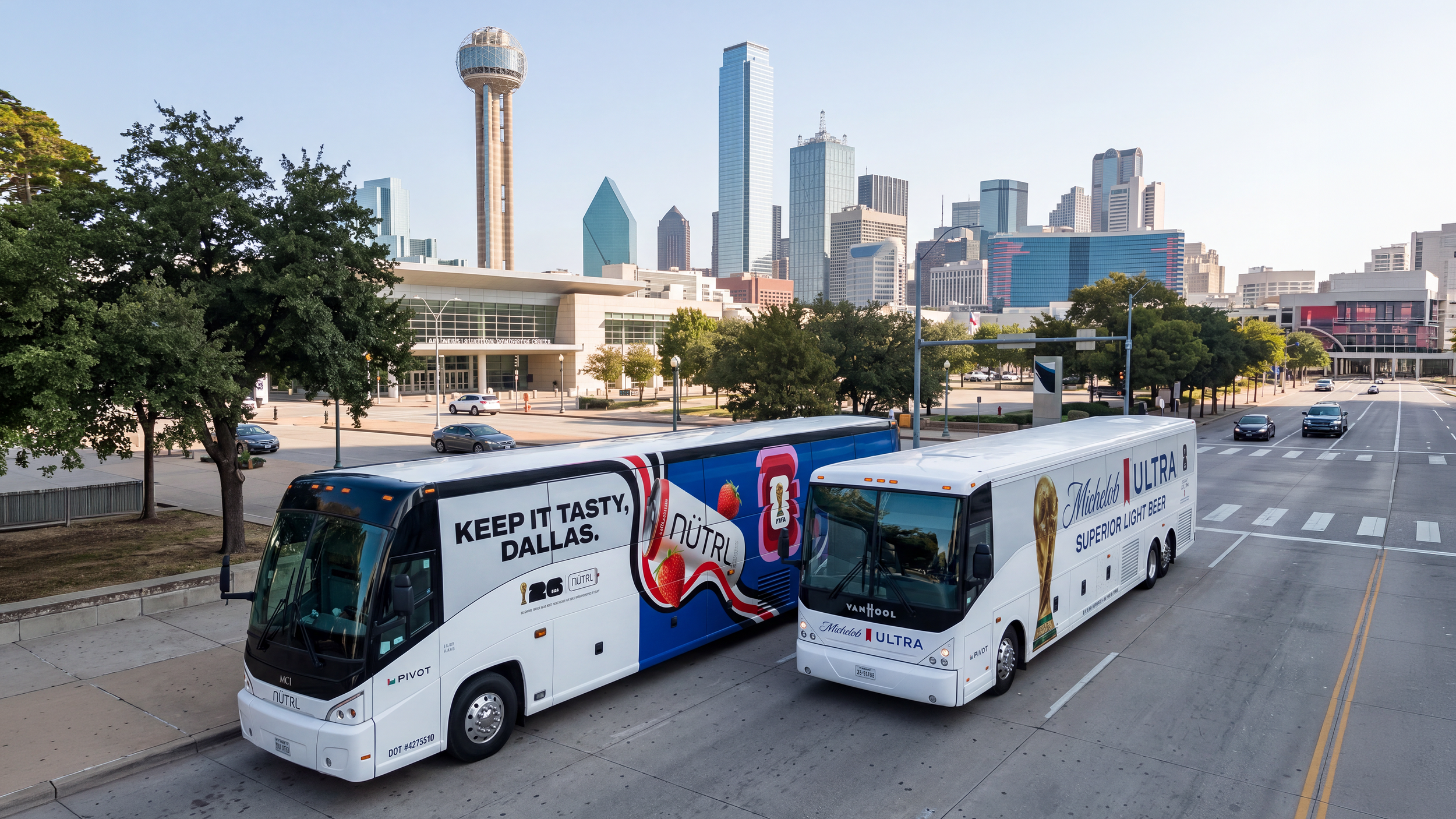Two tour buses parked on a city street with downtown Dallas skyline in the background, including buildings and a tower.