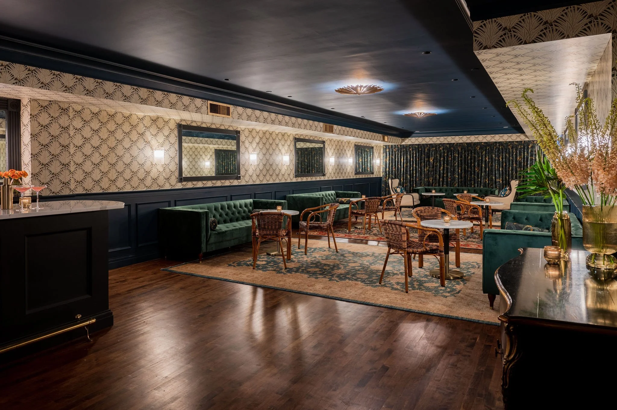 Interior of a stylish lounge or restaurant with dark wood flooring, green velvet sofas, various chairs, and round tables, floral wallpaper, framed mirrors, wall sconces, and ceiling lights, with floral arrangements and curtains.