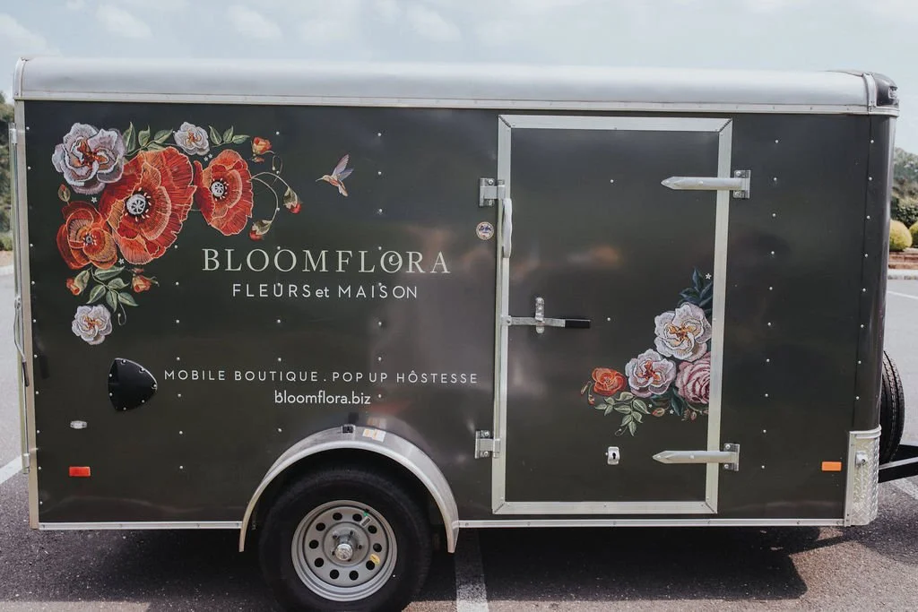A black mobile floral boutique trailer with floral artwork and text for Bloomflora, featuring a door on the right side and a wheel at the bottom