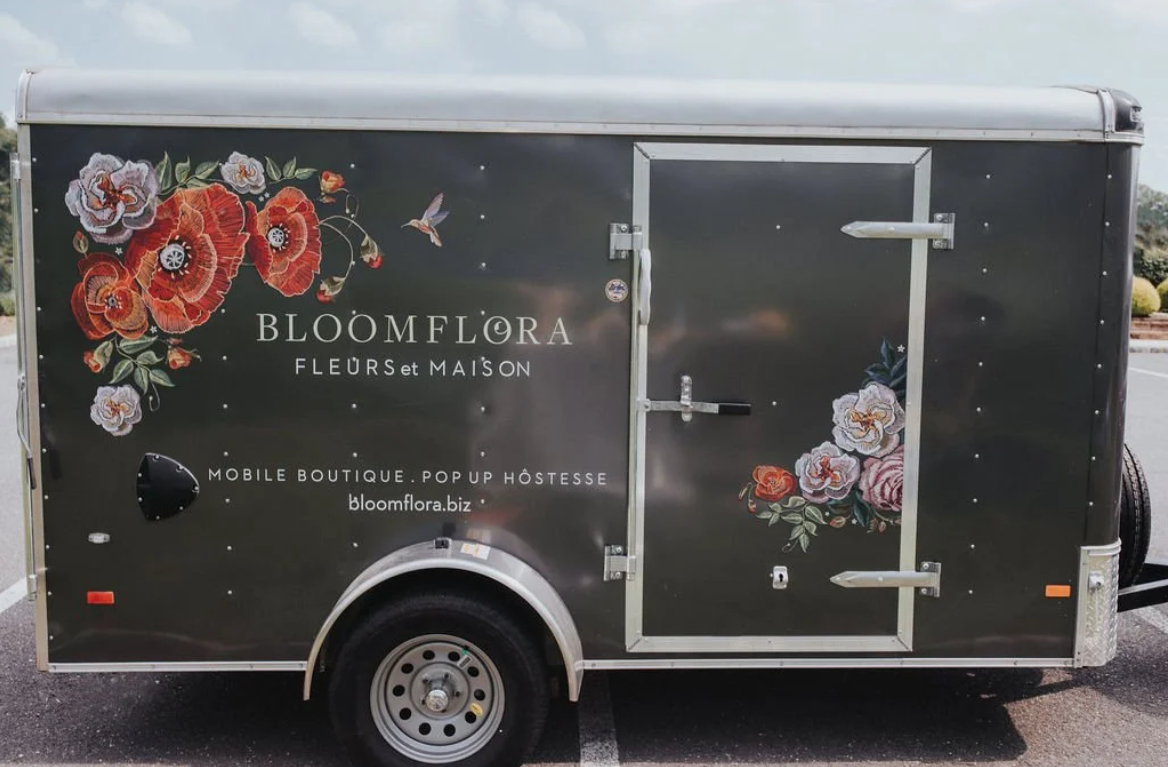 Black mobile boutique trailer decorated with floral artwork and the name Bloomflora, featuring white and red flowers, hummingbird, and information about the business.