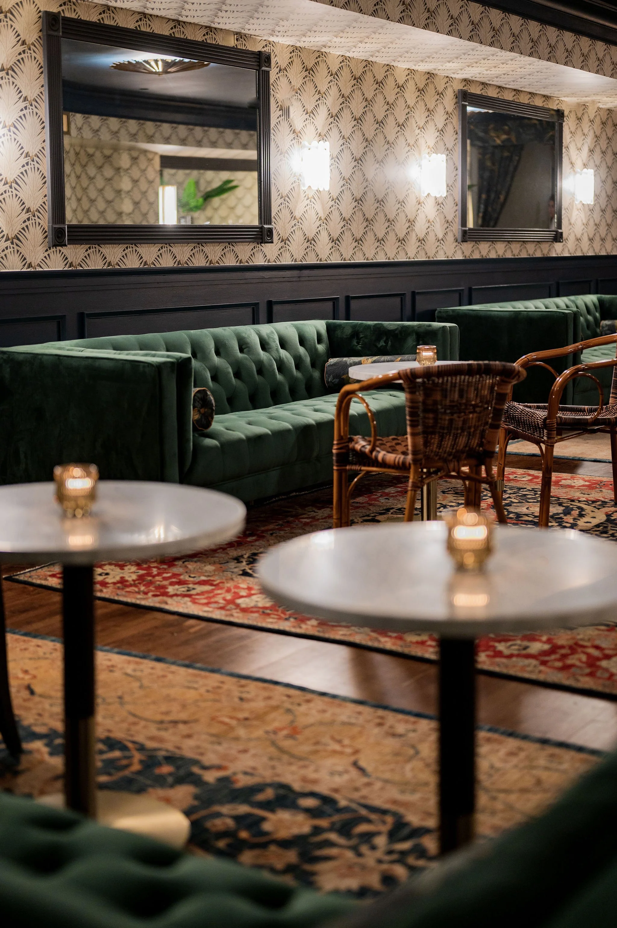 Interior of a cozy lounge area with green velvet tufted sofas, wooden chairs, marble-top tables with small candles, patterned wallpaper, wall-mounted lights, framed mirrors, and a colorful rug.