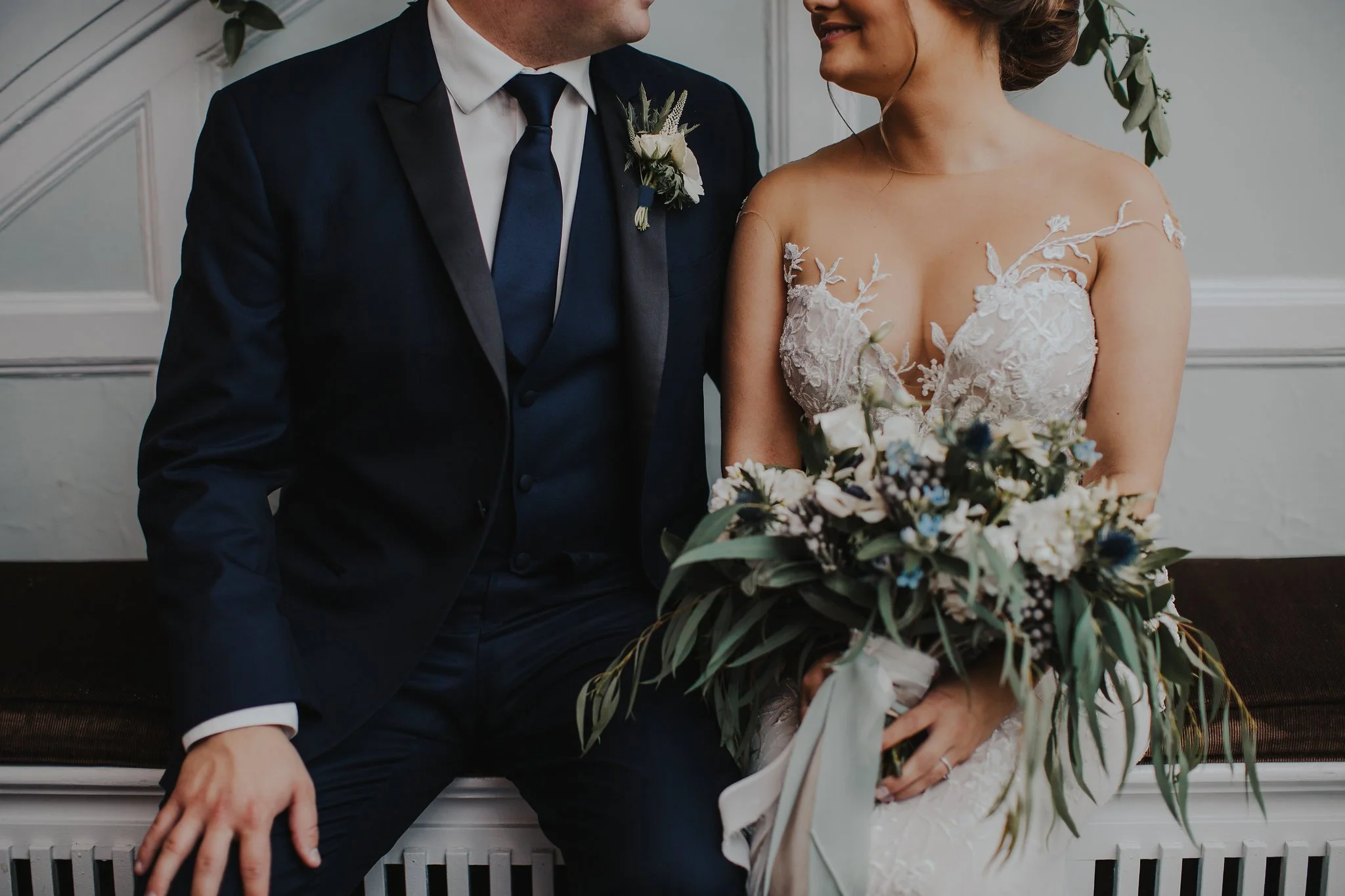 Close-up of a bride and groom sitting together, smiling, during their wedding. The bride wears an off-the-shoulder lace wedding gown, and the groom wears a navy blue suit with a white shirt and tie. They are holding a bouquet of flowers, and the background features a light-colored wall with decorative molding.