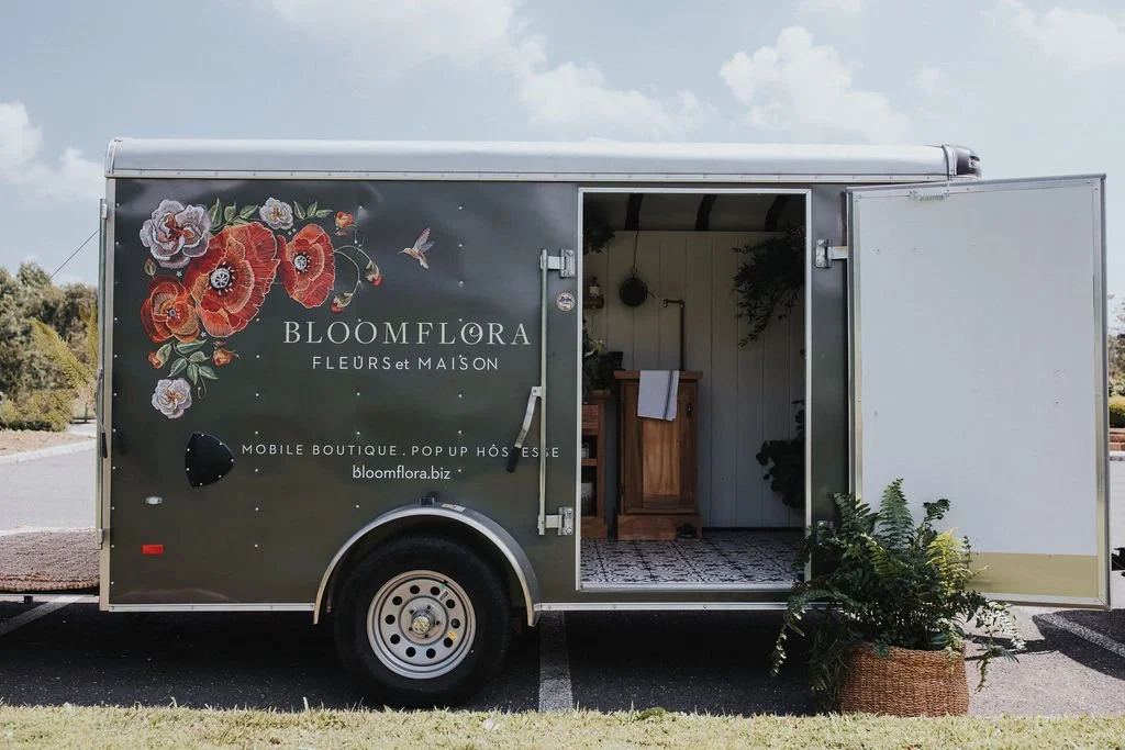 A mobile floral boutique trailer with closed sides, one open door, floral and hummingbird graphics, and a potted plant outside