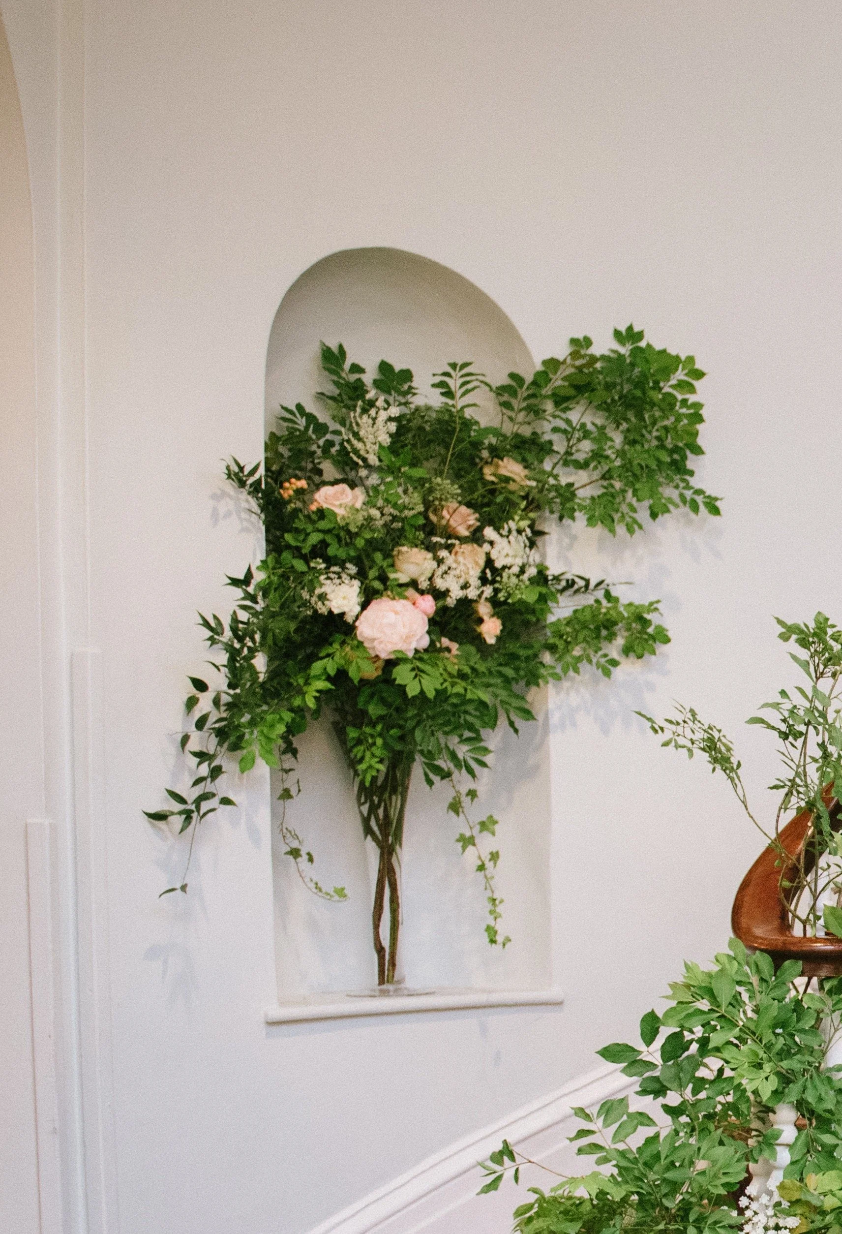 A floral arrangement with pink and white flowers and green foliage in front of a white wall and arched wall niche.