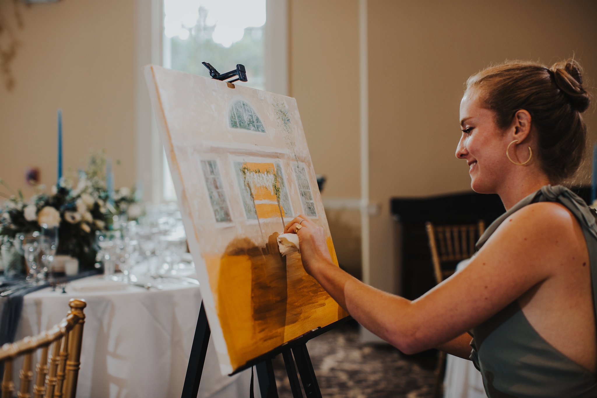 A woman painting a large watercolor or acrylic landscape on a canvas set on an easel indoors.