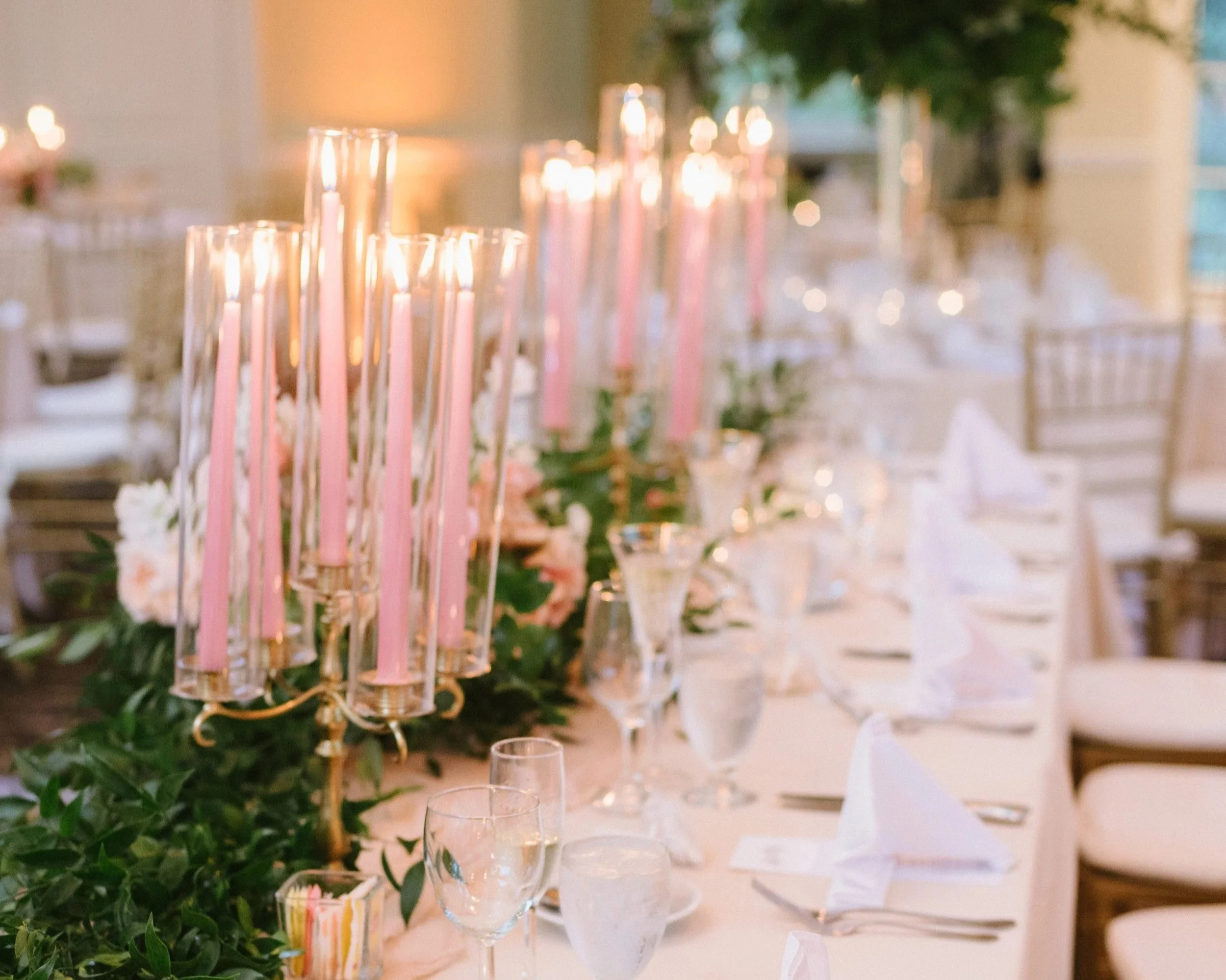 Elegant table setting with tall pink candles in glass holders, surrounded by greenery and glassware, likely for a wedding or formal event.