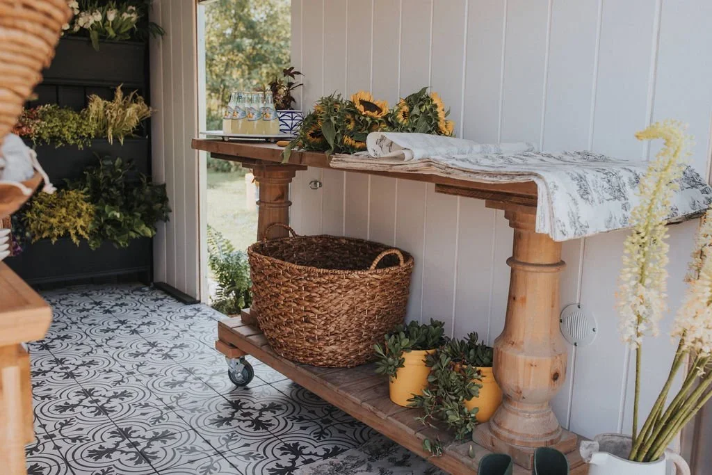 Indoor space with a wooden console table holding a sunflower arrangement, rolled cloth, and glass bottles, against a white wall with a window, tiled patterned floor, and plants in baskets and pots.