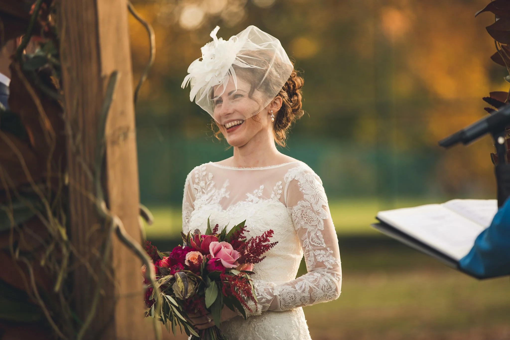 A smiling woman in a lace wedding dress holding a bouquet of pink and red flowers during a ceremony outdoors at sunset.