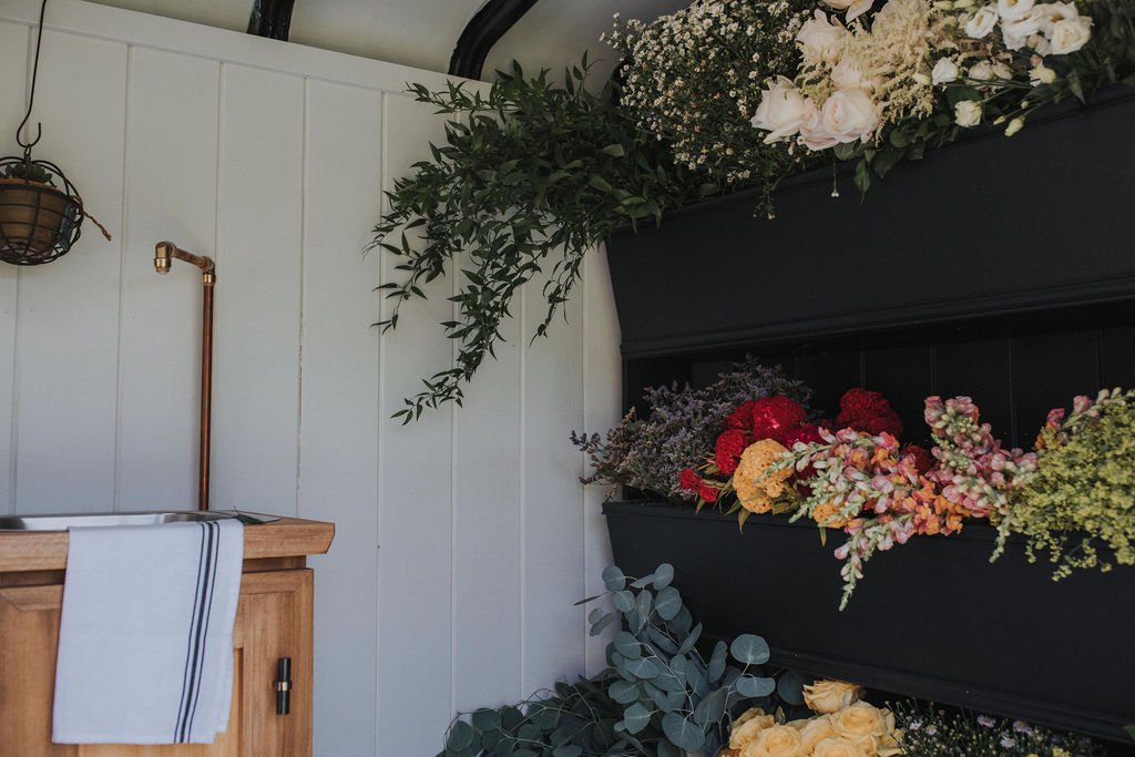 Indoor floral display with various flowers, greenery, and a wooden cabinet with a towel hanging on it.